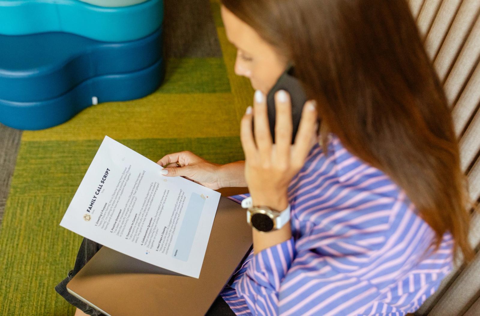 A woman sits and talks on a cell phone. She is holding a piece of paper in front of her.