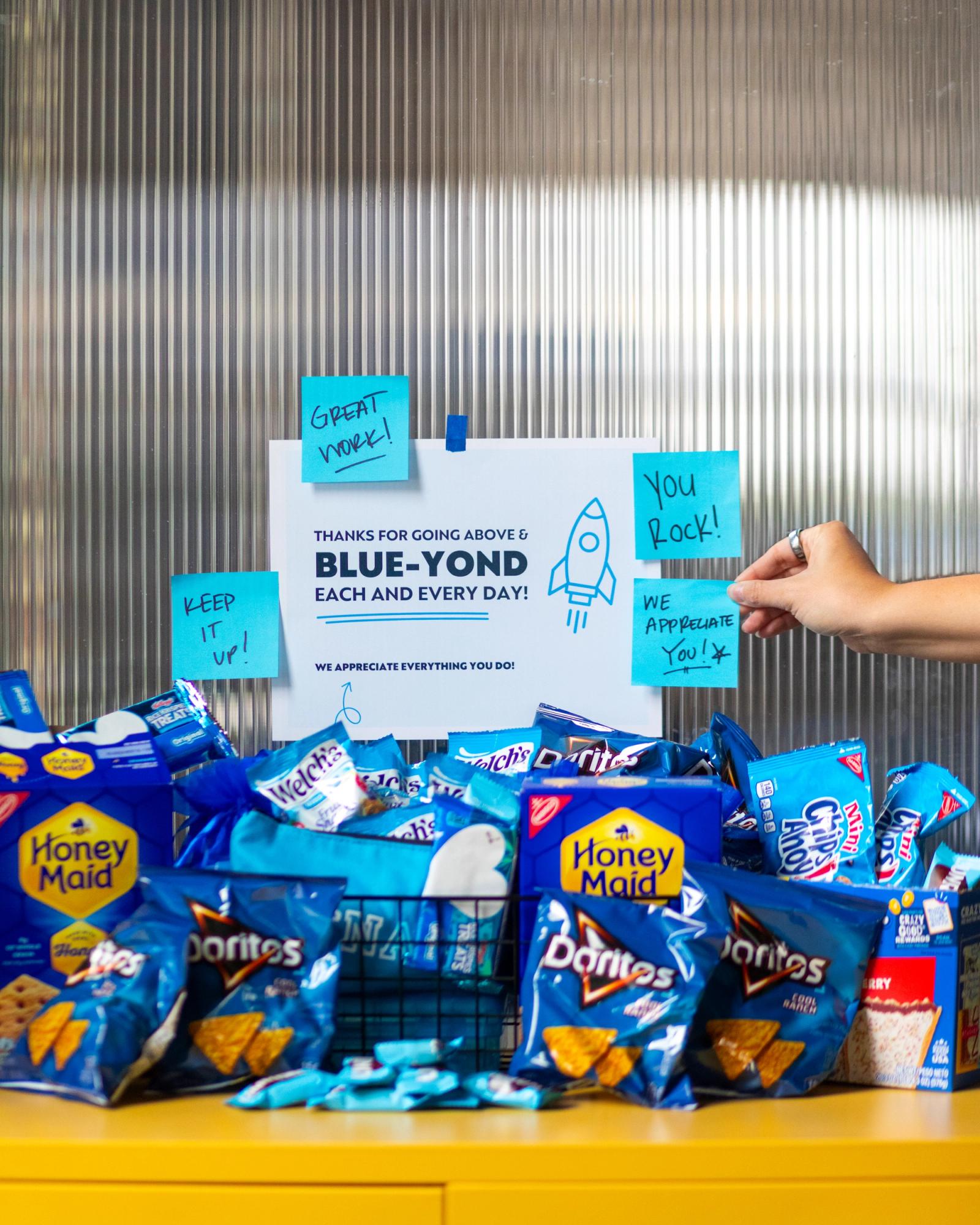 Small snacks in blue bags sit on a table in front of a glass wall. A poster and blue sticky notes are on the wall.