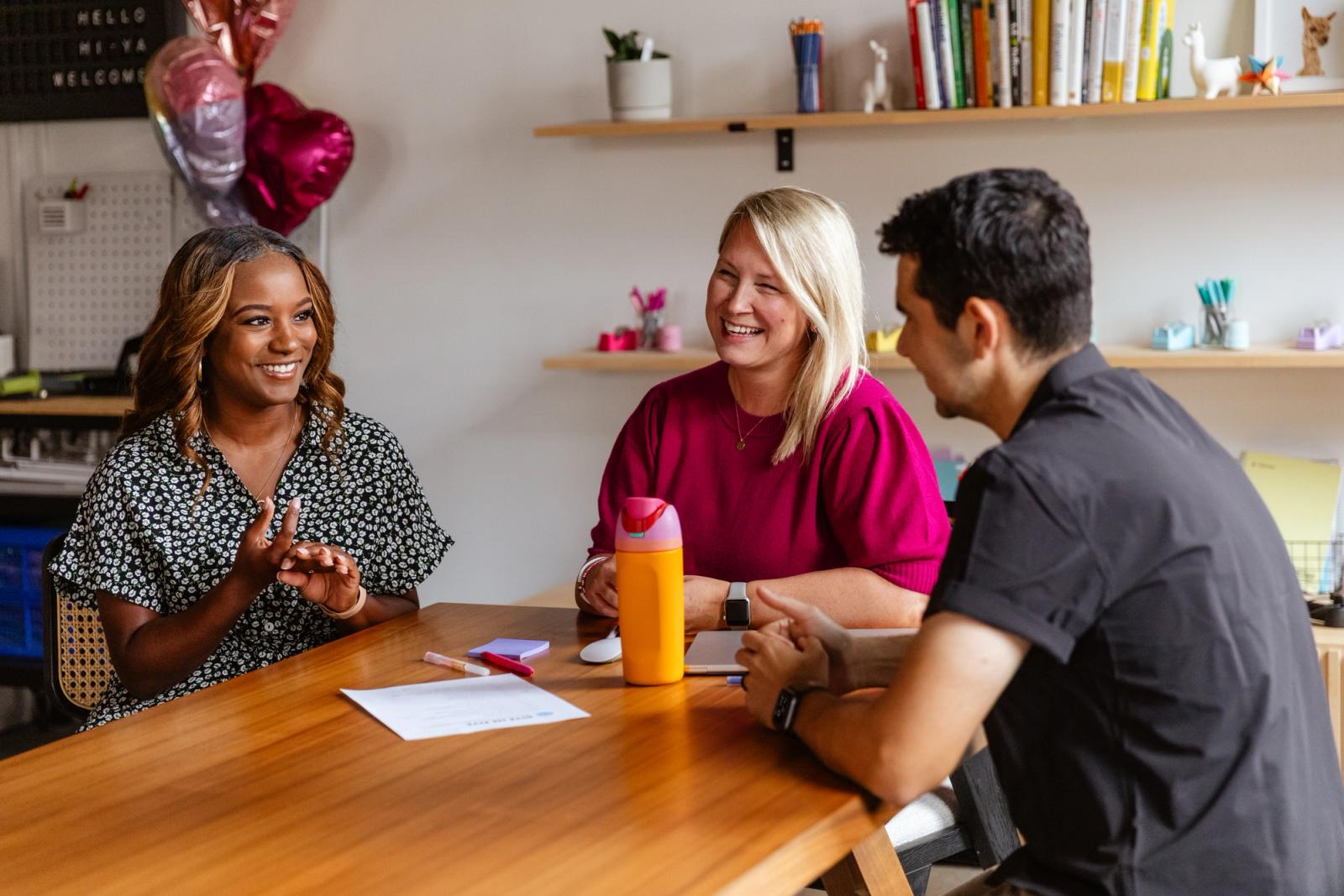 Three people sit at a table laughing and smiling at each other. 