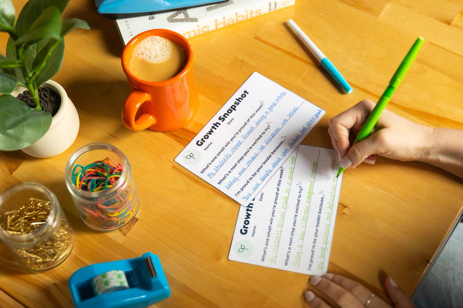 A hand writes with a green pen on a small postcard on a desk filled with office supplies, a cup of coffee, and small plant.