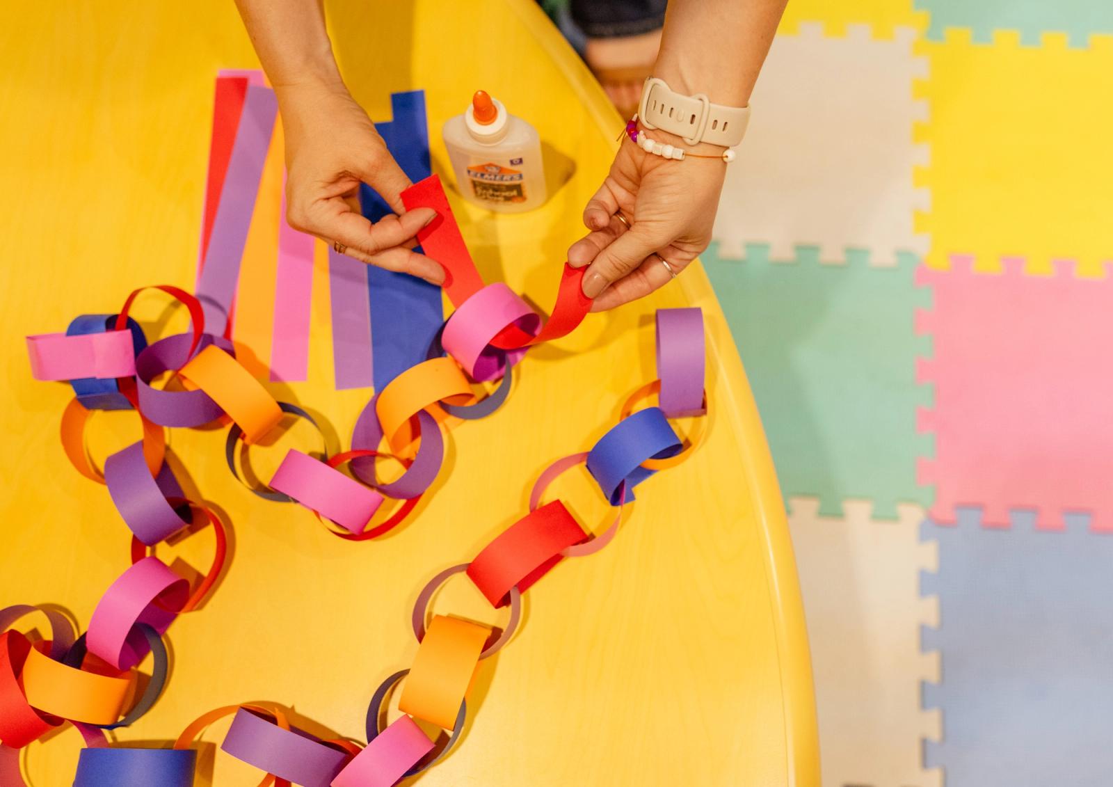 A colorful paper chain lays on a yellow table. Two hands reach in to add another link to the chain.