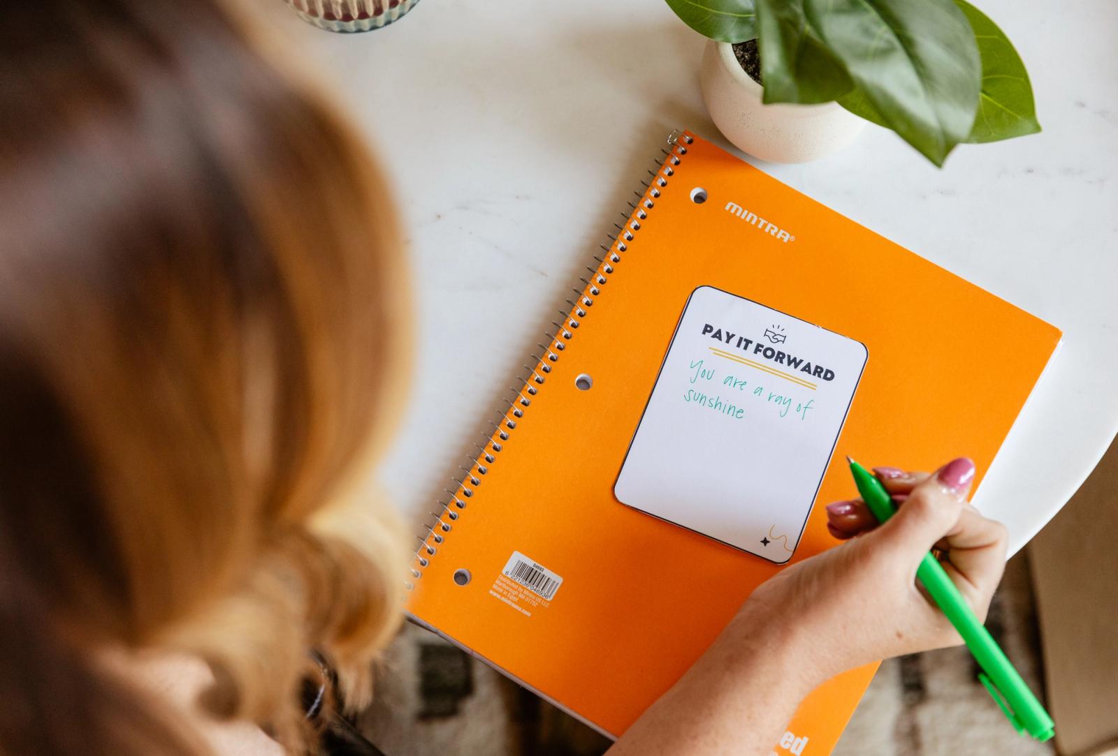 A person writes on a notecard that says "pay it forward" on top of an orange notebook.
