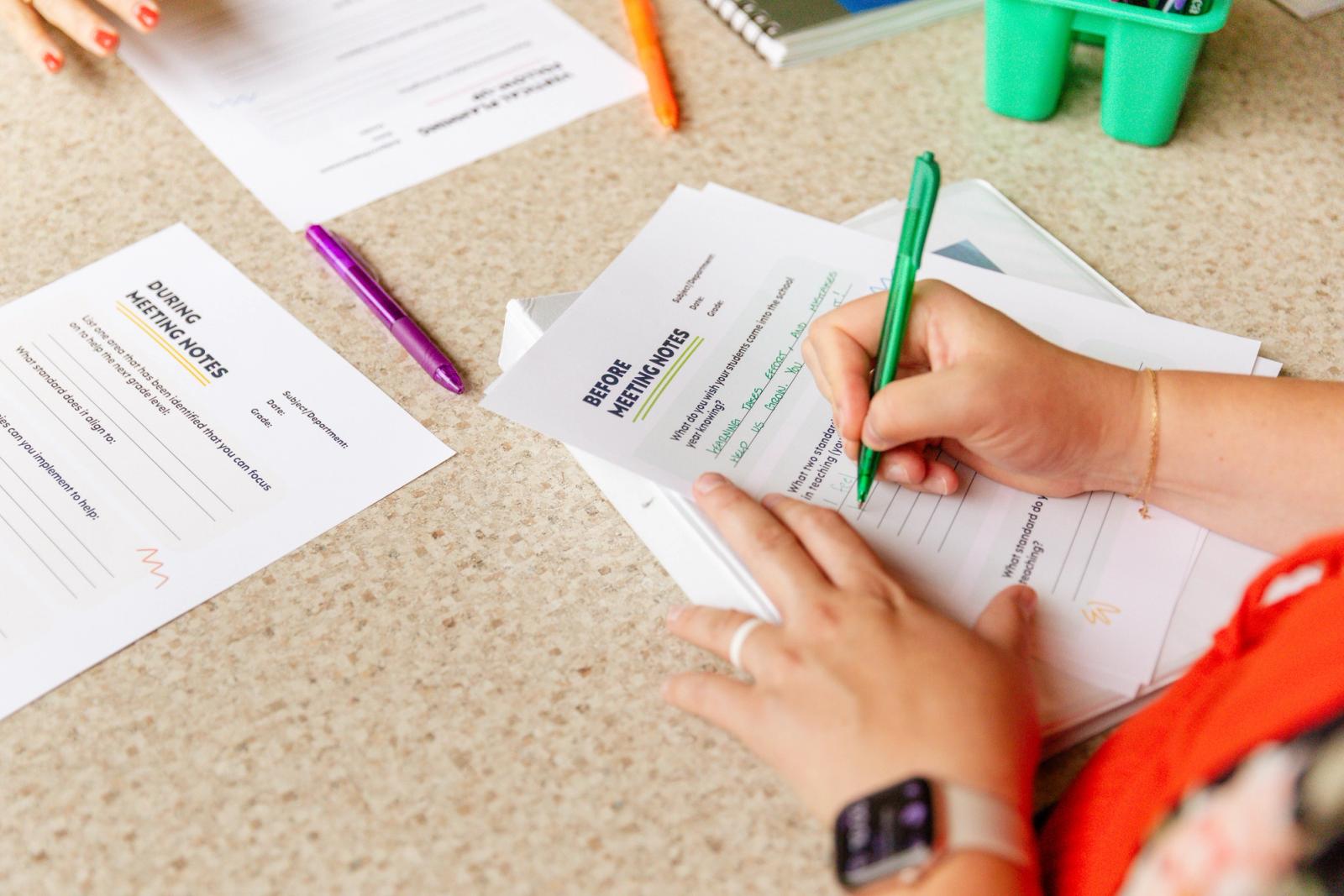 A woman sits at a table with papers and pens scattered on it. She writes with a green pen on a piece of paper.