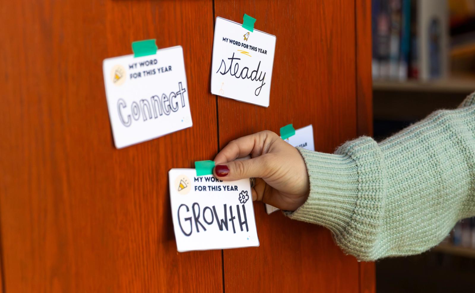 Three notecards each with one word on them are taped to a red wood wall. A hand reaches in from the side to tape one more notecard on the wall.