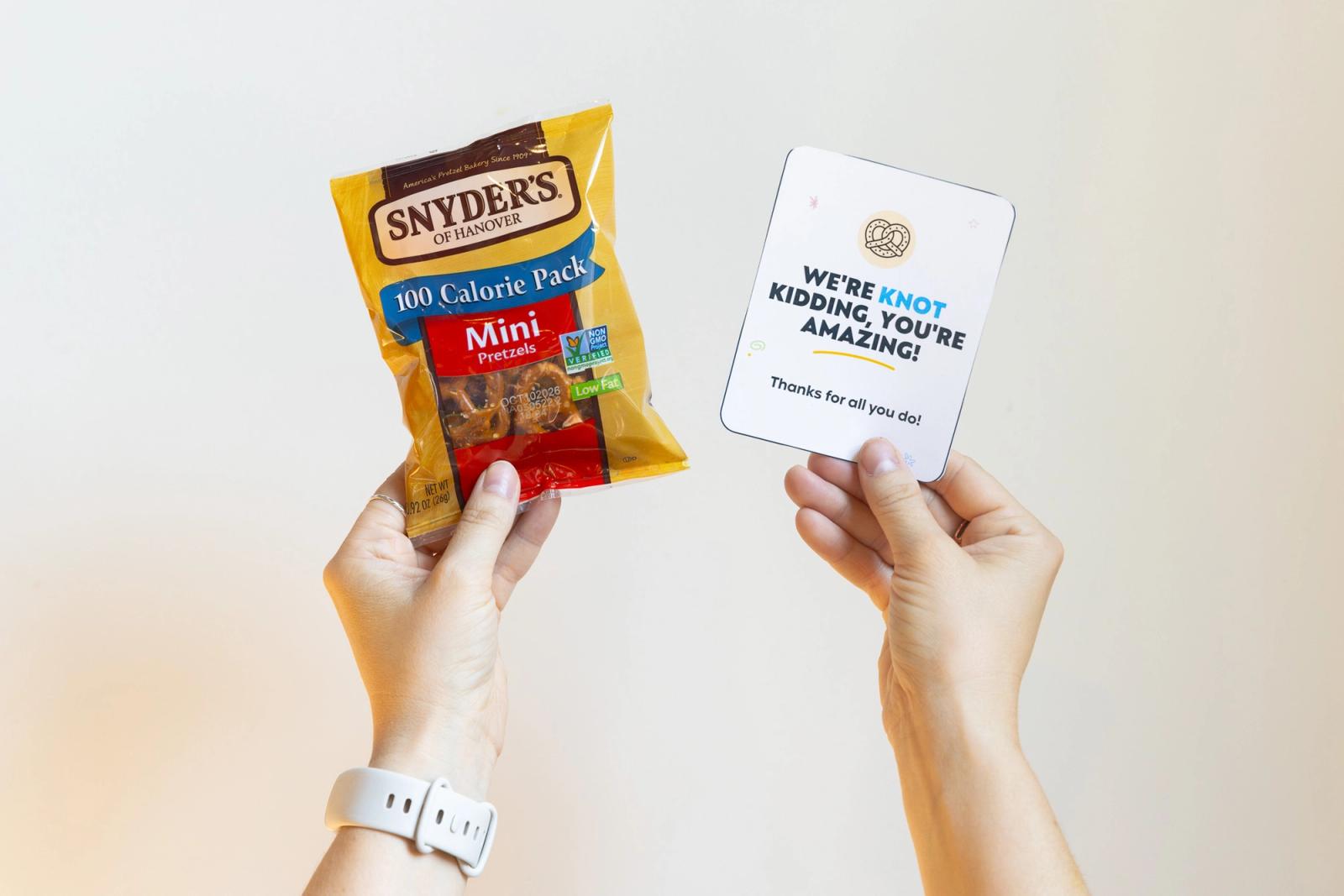 Hands hold up a small bag of pretzels and a small piece of paper in front of a white wall.