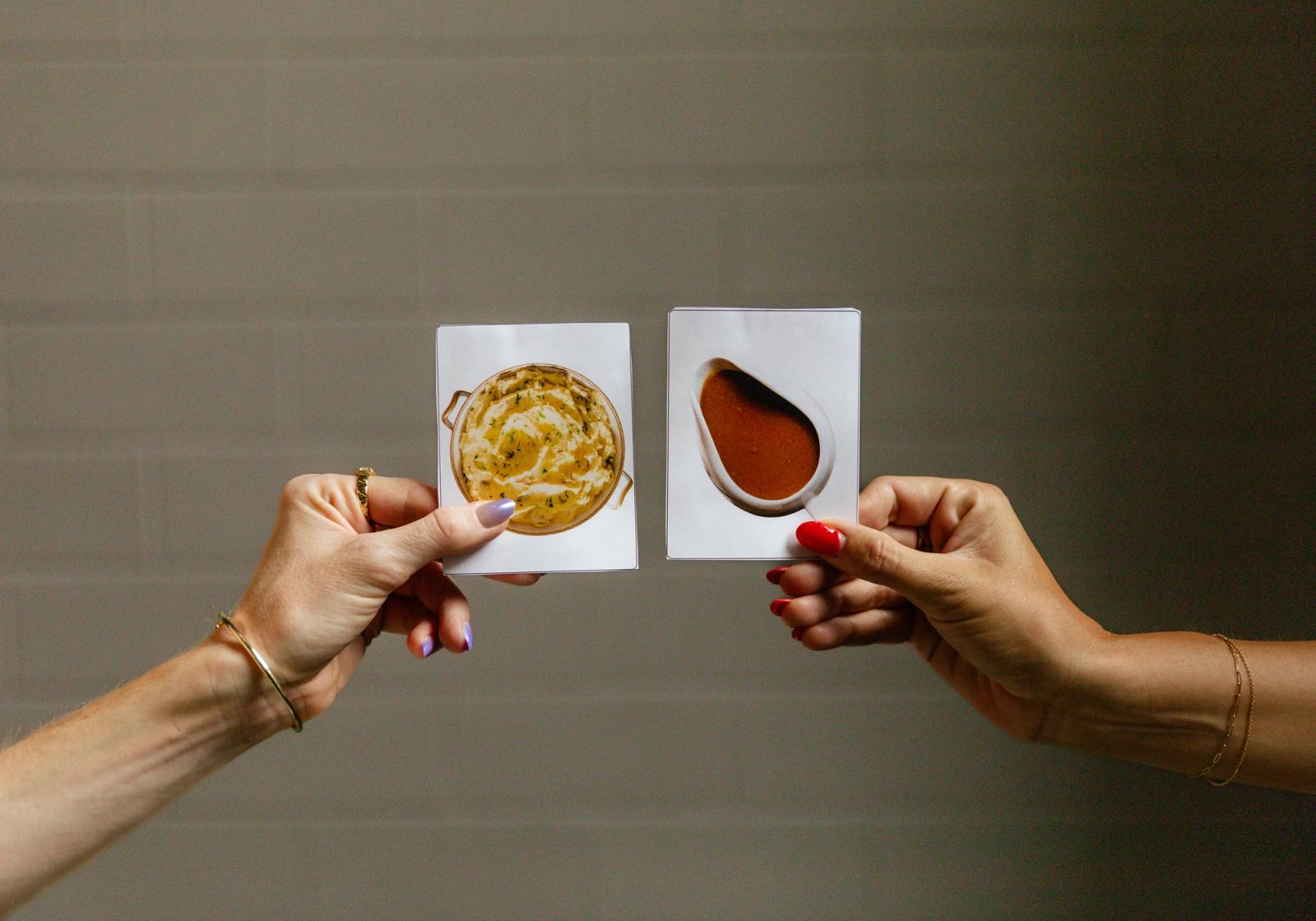 Two hands reach in from the side each holding a piece of paper, one with a picture of mashed potatoes, the other with a picture of gravy.