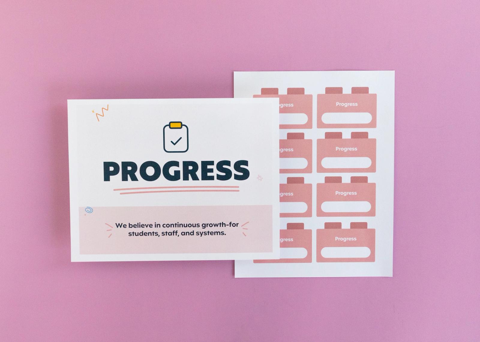 Sheet of paper that says "progress" and another with small squares on it lay on a pink background.