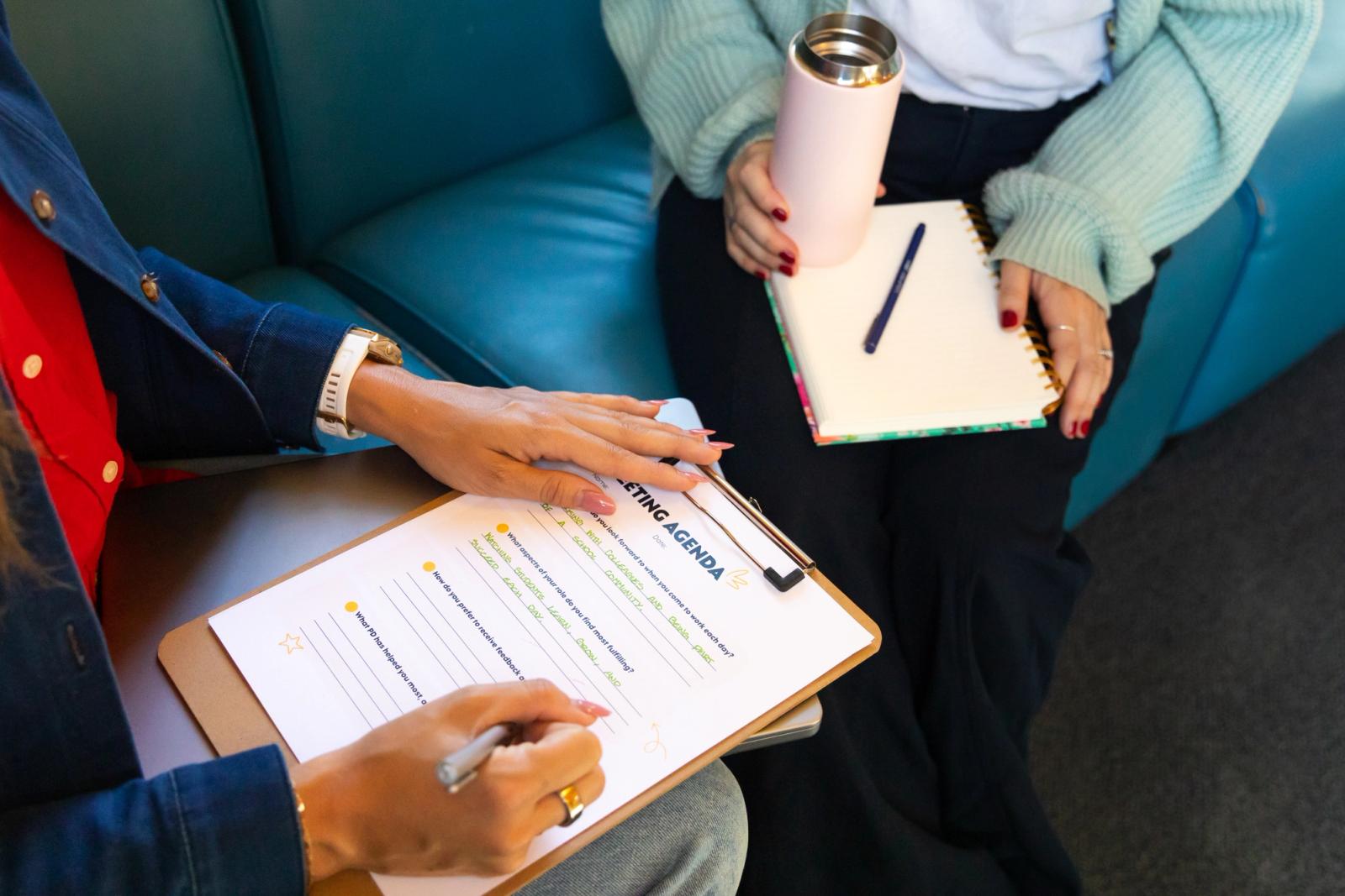 A woman writes on a piece of paper on a clipboard. Another women sits across from her with a notebook, pen, and coffee cup.