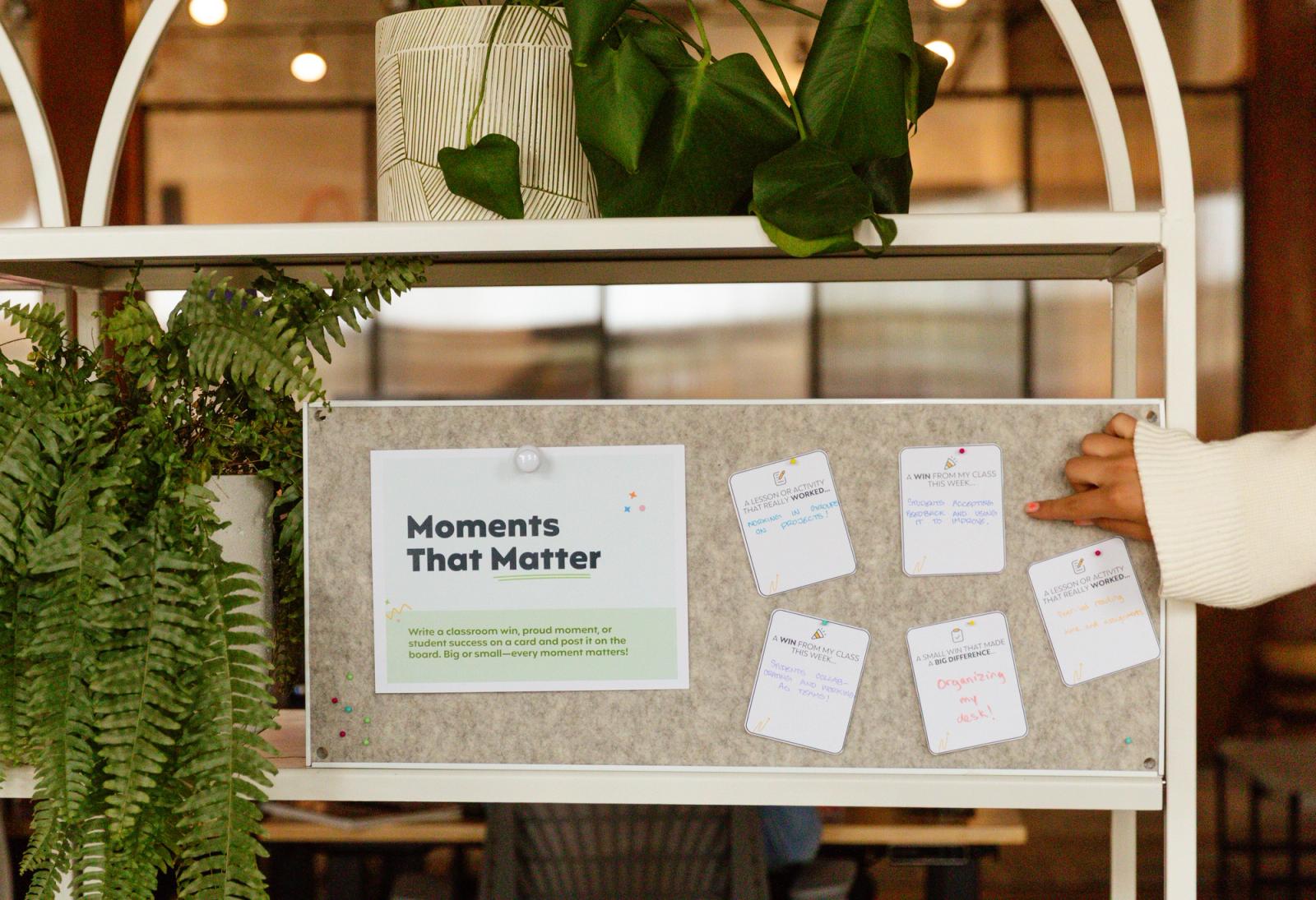 A bulletin board with a poster that says "moments that matter" on it and small notecards sits on a shelf next to two potted plants.