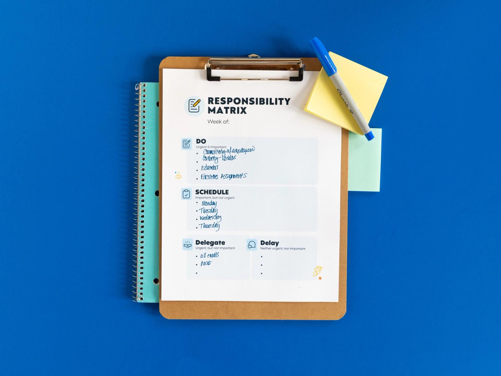 A clipboard with a sheet of paper that says "responsibility matrix" on it sits on a notebook, all on a blue background. Sticky notes and a pen are on top of the clipboard.