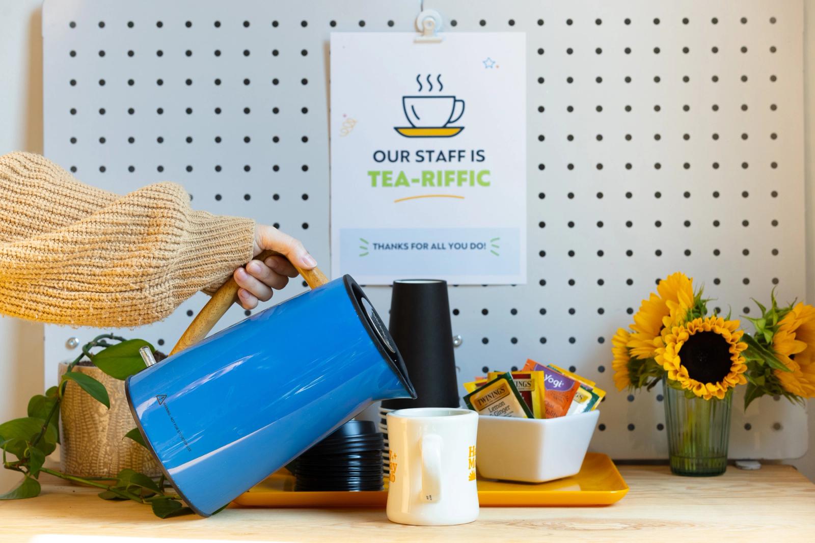 A tea station with flowers is set up on a table. A hand reaches in and pours water into a mug.
