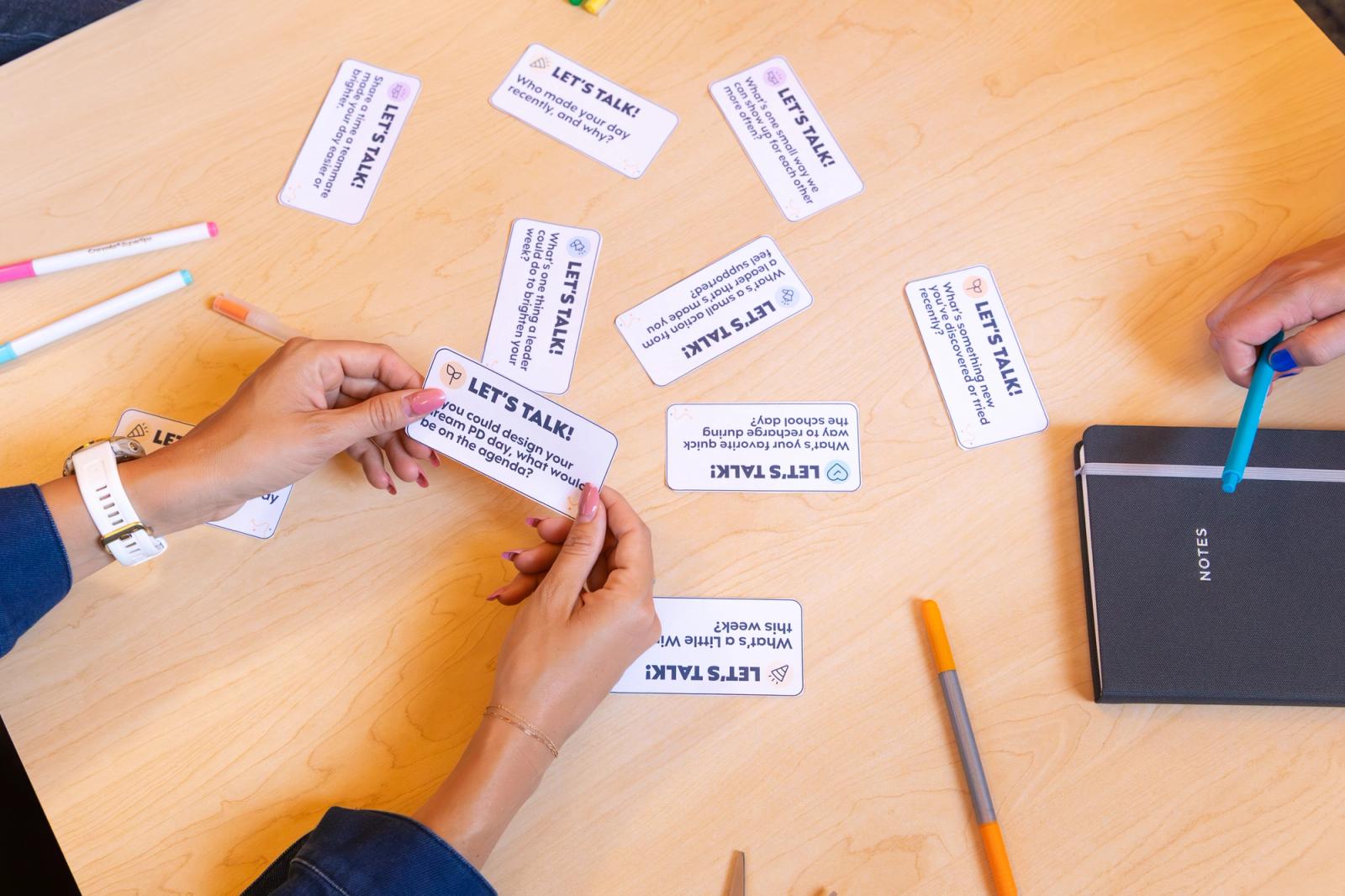Hands hold a small piece of paper that says "let's talk" on it. Several other small notecards lay on a table with notebooks and pens.