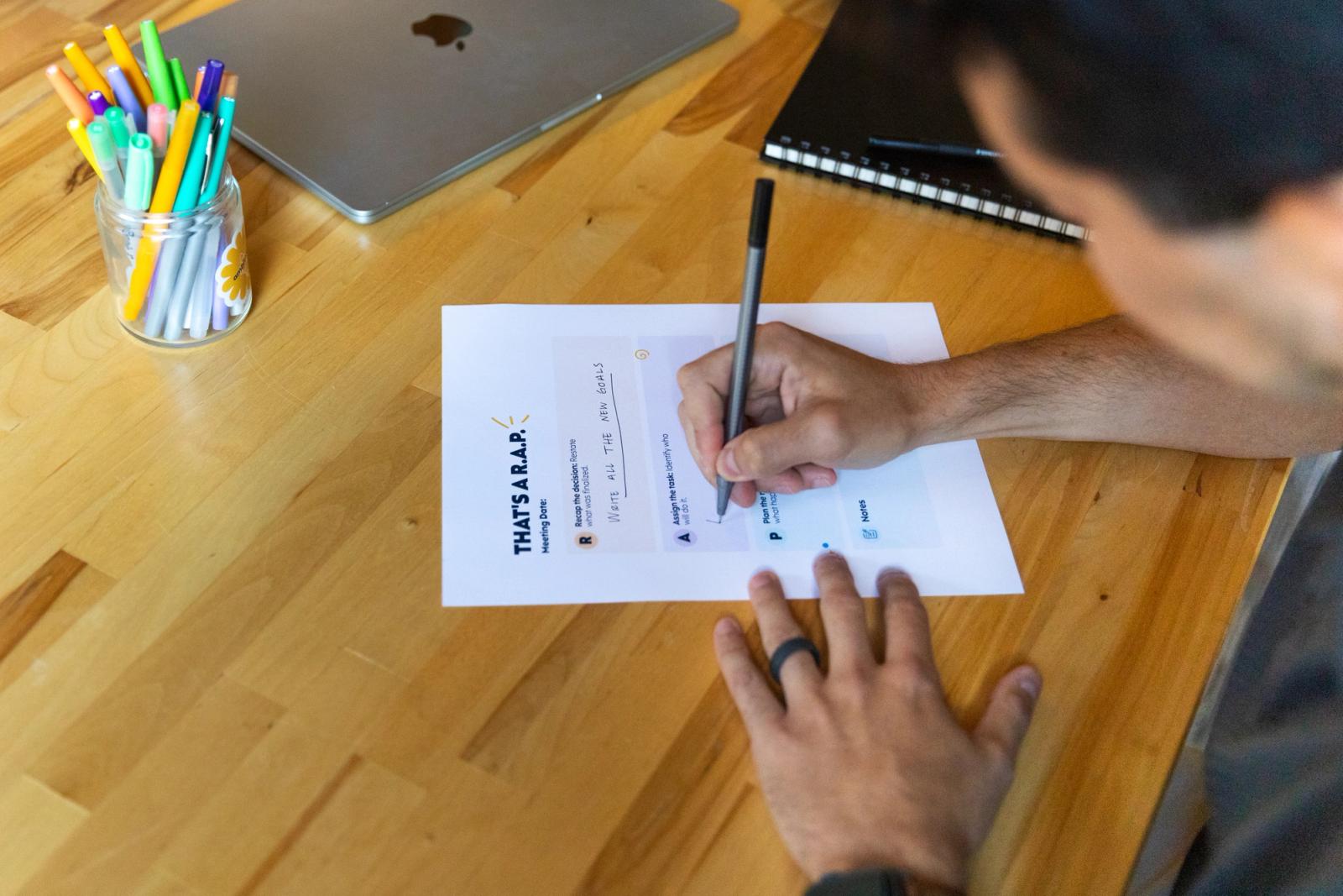 A man writes on a piece of paper on a table with a jar of pens and laptop on it. 