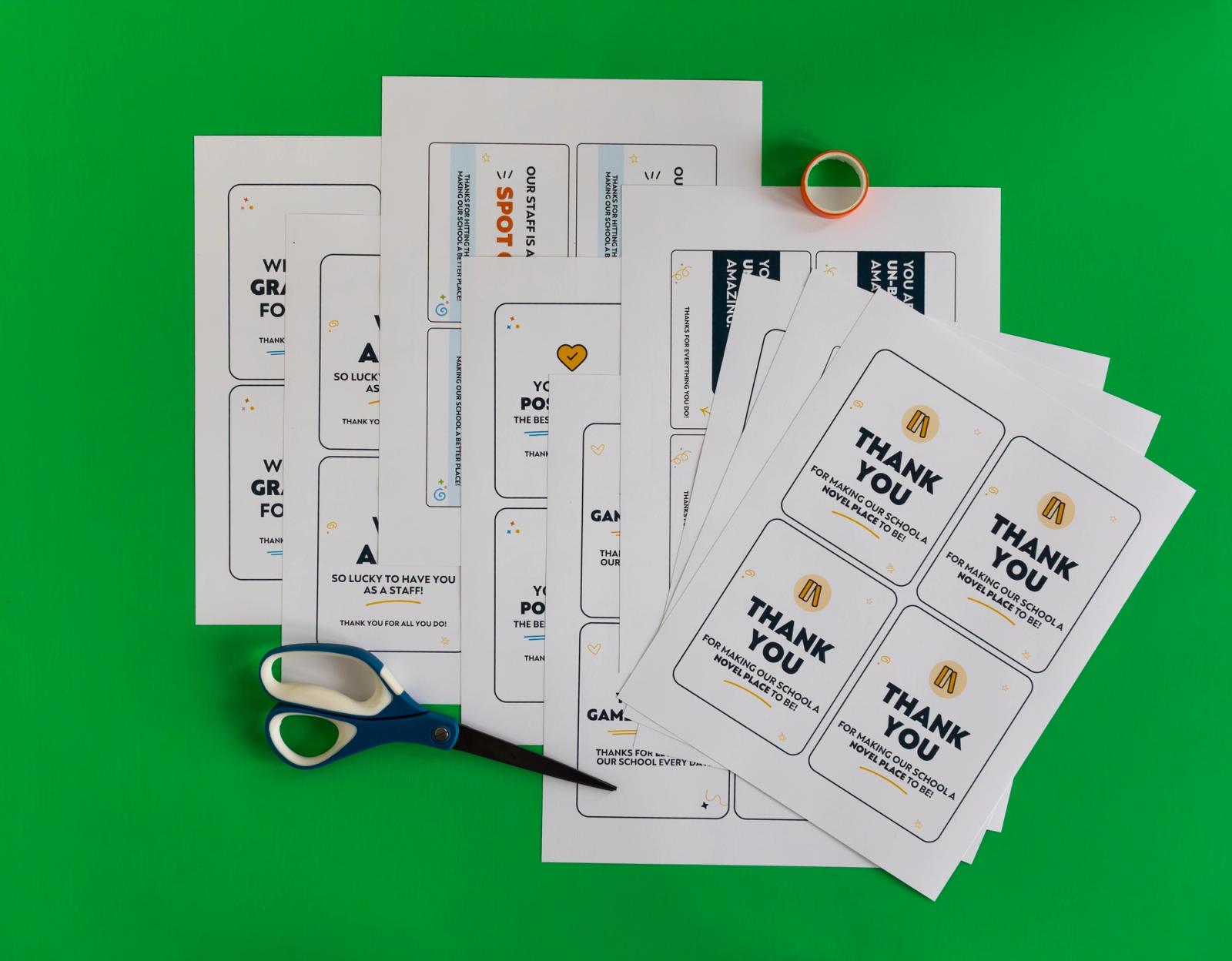 Several sheets of paper with notecard outlines on them lay on a green background. A pair of scissors and roll of tape lay to the side.