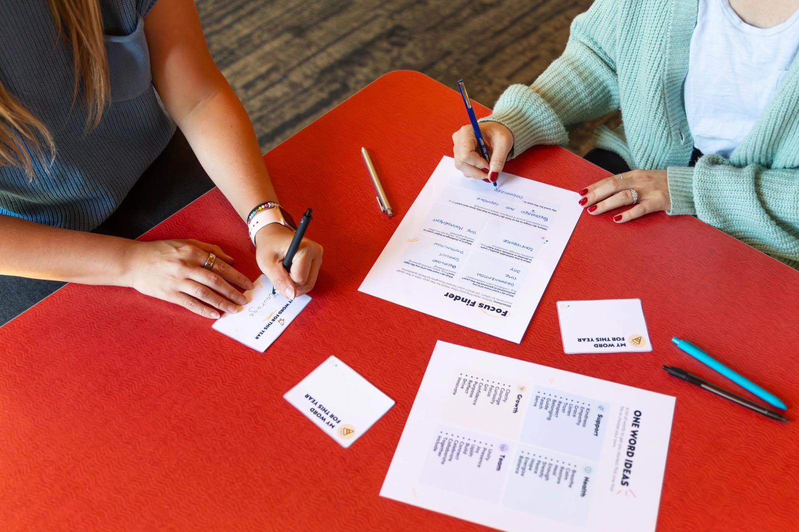 Two people sit at a red table writing on pieces of paper with pens and more paper scattered on the table.
