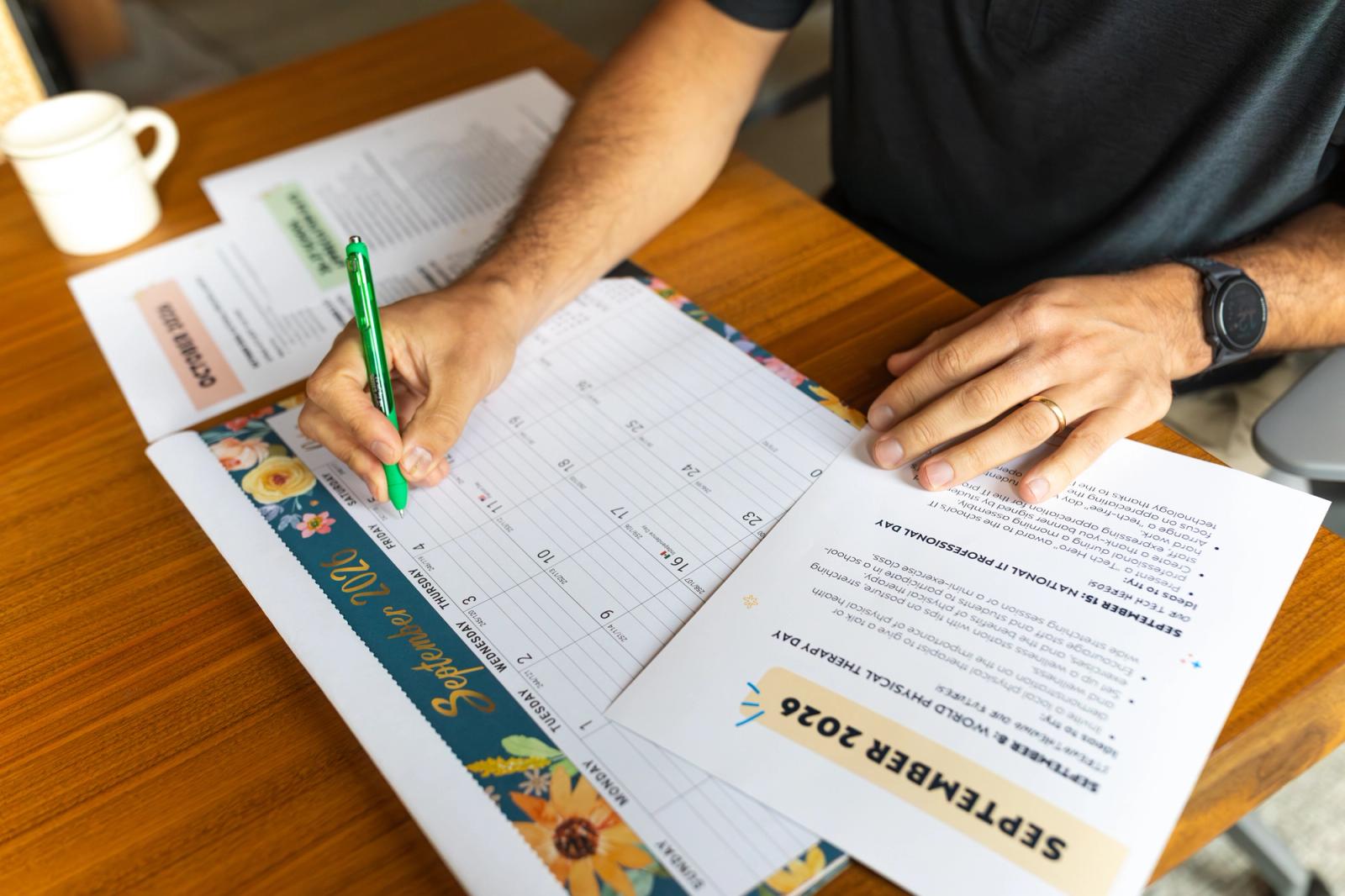 A man sits as a desk with a September calendar in front of him. He holds a green pen and marks the calendar.