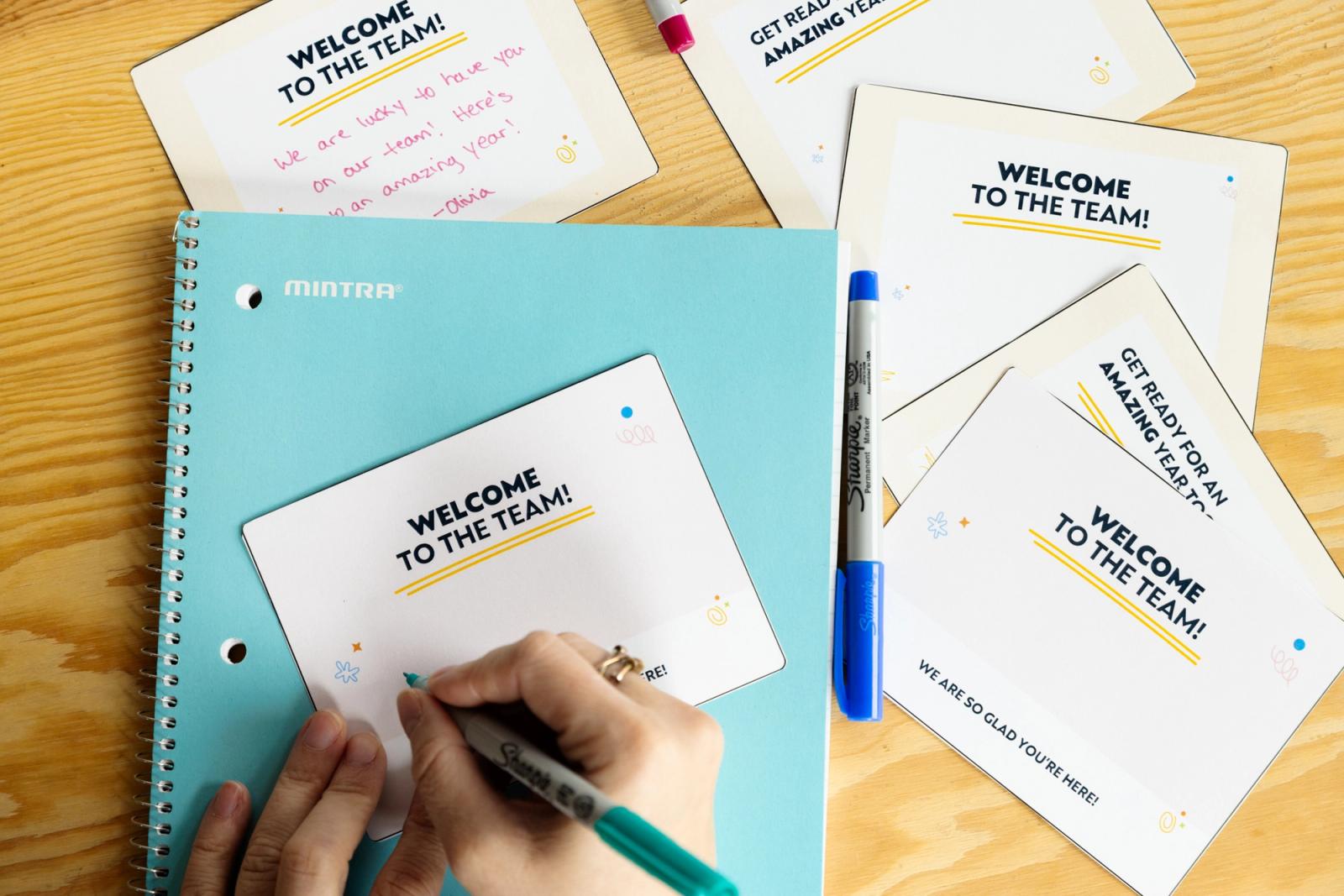 A hand writes on a postcard that says "welcome to the team." 