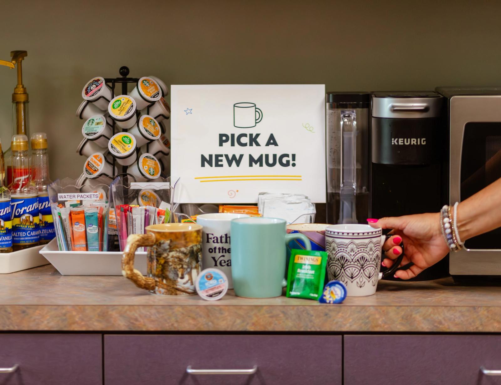 A coffee maker, coffee supplies, and colorful mugs sit on a counter. A hand reaches in to grab a mug.