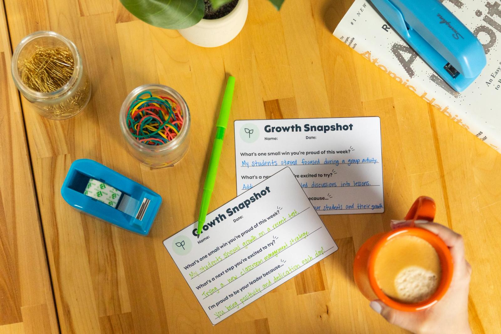Two postcards lay on a wooden desk with a green pen, coffee cup, and office supplies.