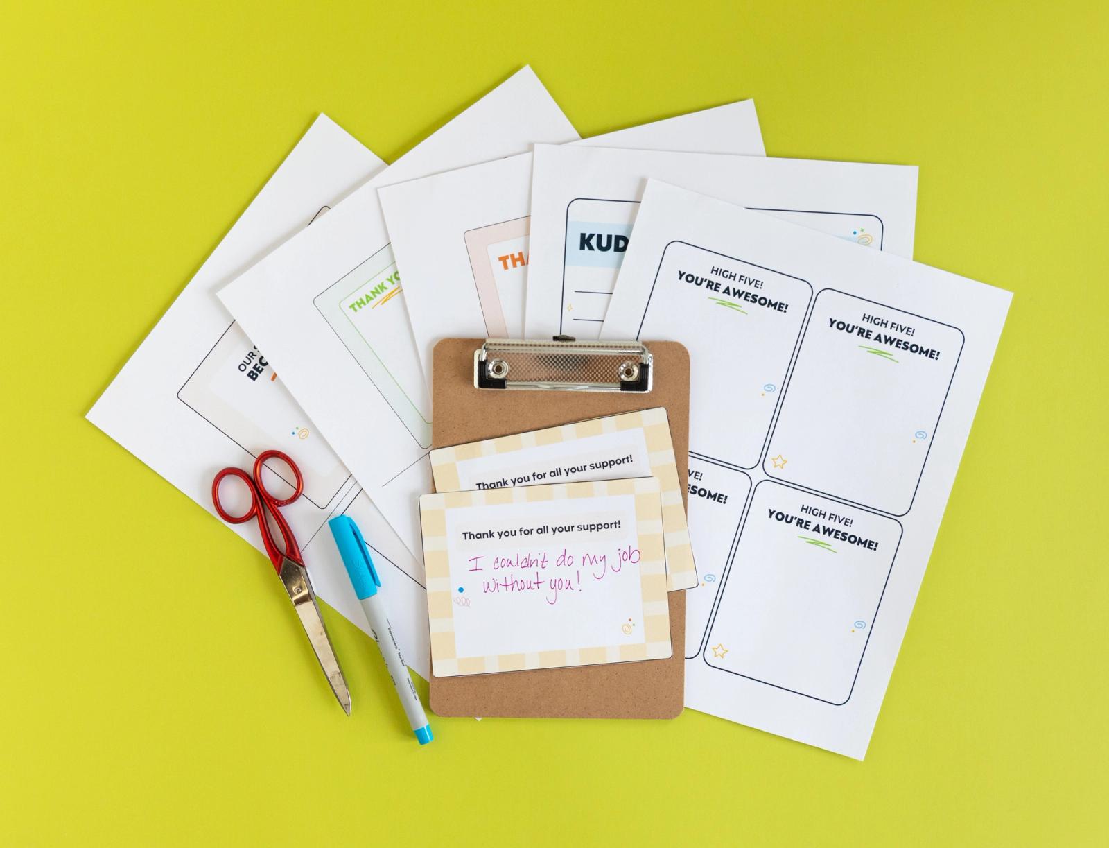 Several sheets of paper, fanned out, lay on light green background. A clipboard with postcards, a blue pen, and pair of scissors lay on top of the papers.