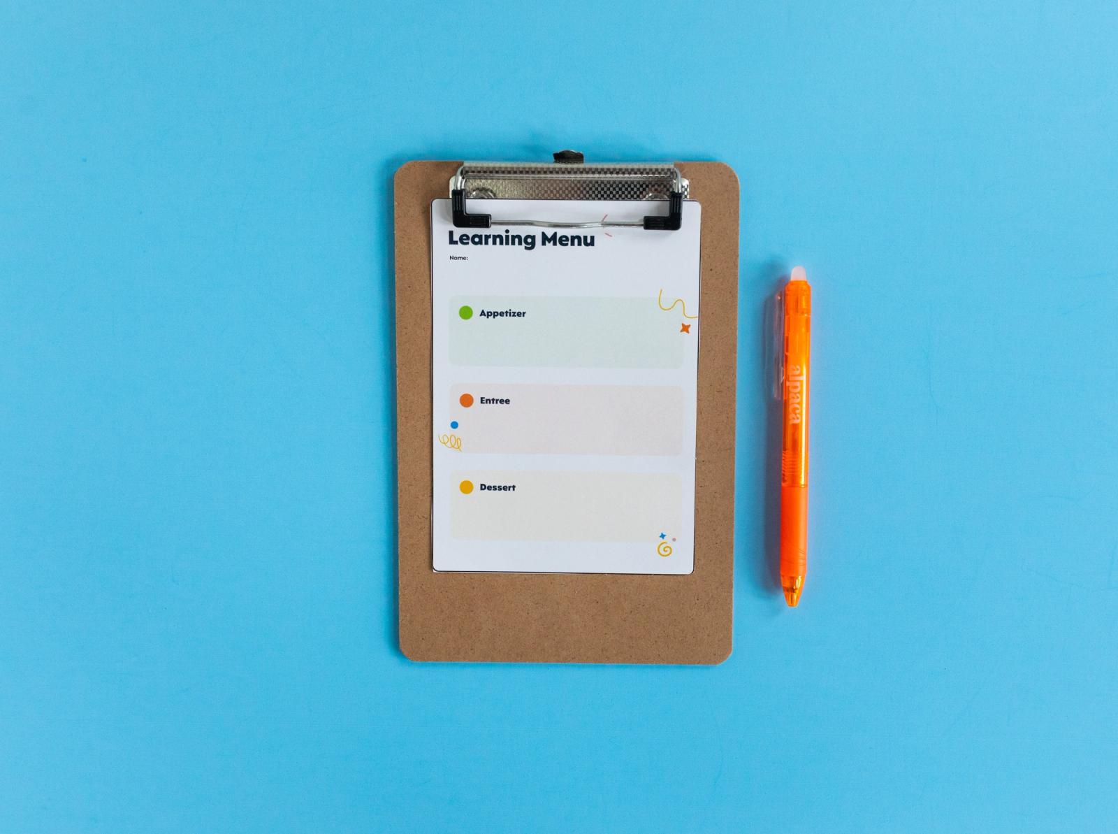 A small clipboard with a piece of paper on it that says "learning menu" lays on a light blue background. An orange pen lays beside the clipboard. 