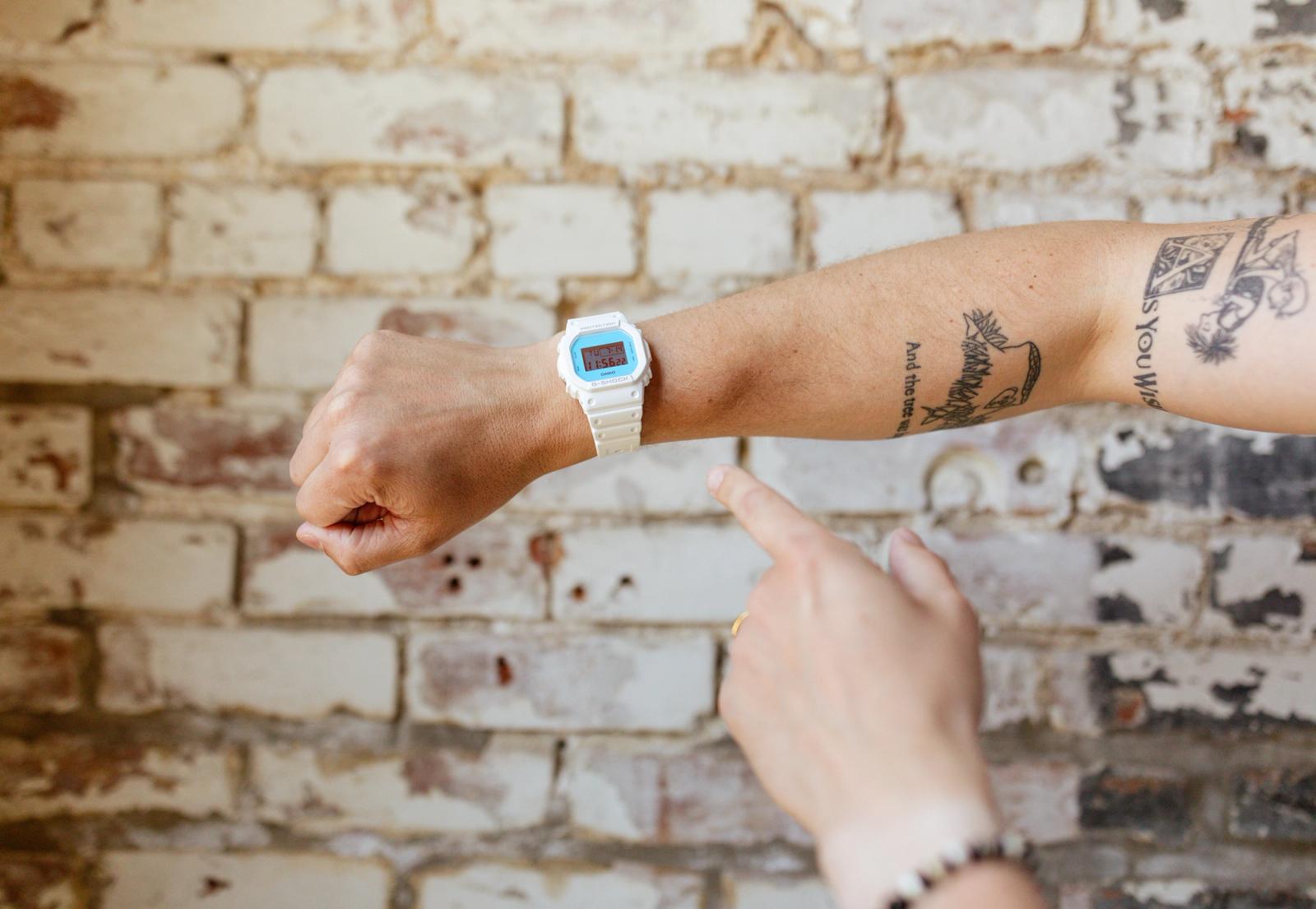 An arm with a white wristwatch reaches in from the left side and a hands points to the watch in front of a white brick background.