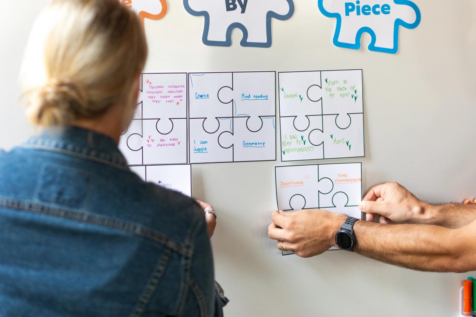 A woman with her back to the camera stands in front of a whiteboard with large paper puzzle pieces taped to it. A man reaches in from the right to place a piece on the whiteboard.