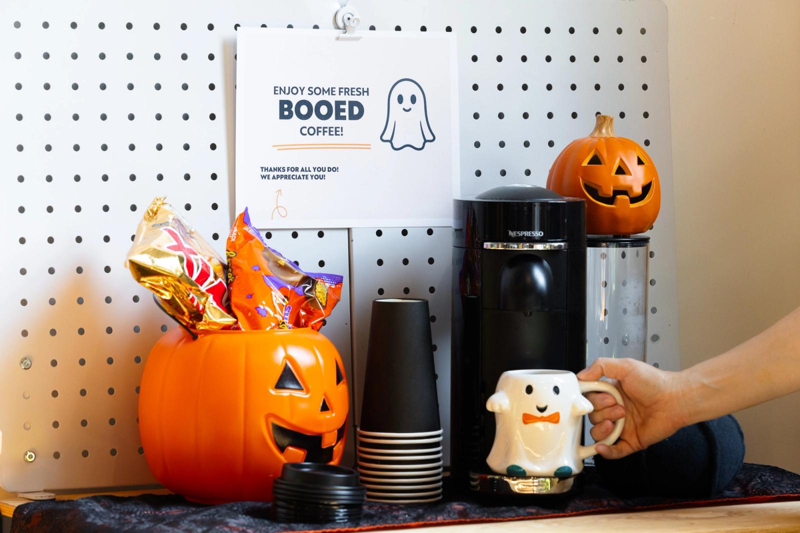 A coffee make, to-go cups, and a pumpkin bucket with candy in it lay on lays on a table. A poster hangs behind it, and a hand reaches in to grab a ghost coffee mug.