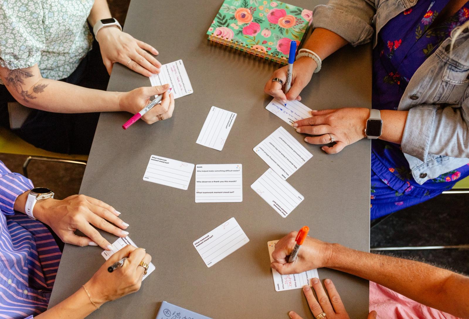 Four people sit at a table. On the table are small notecards scattered. Each of the people write on one of the postcards.