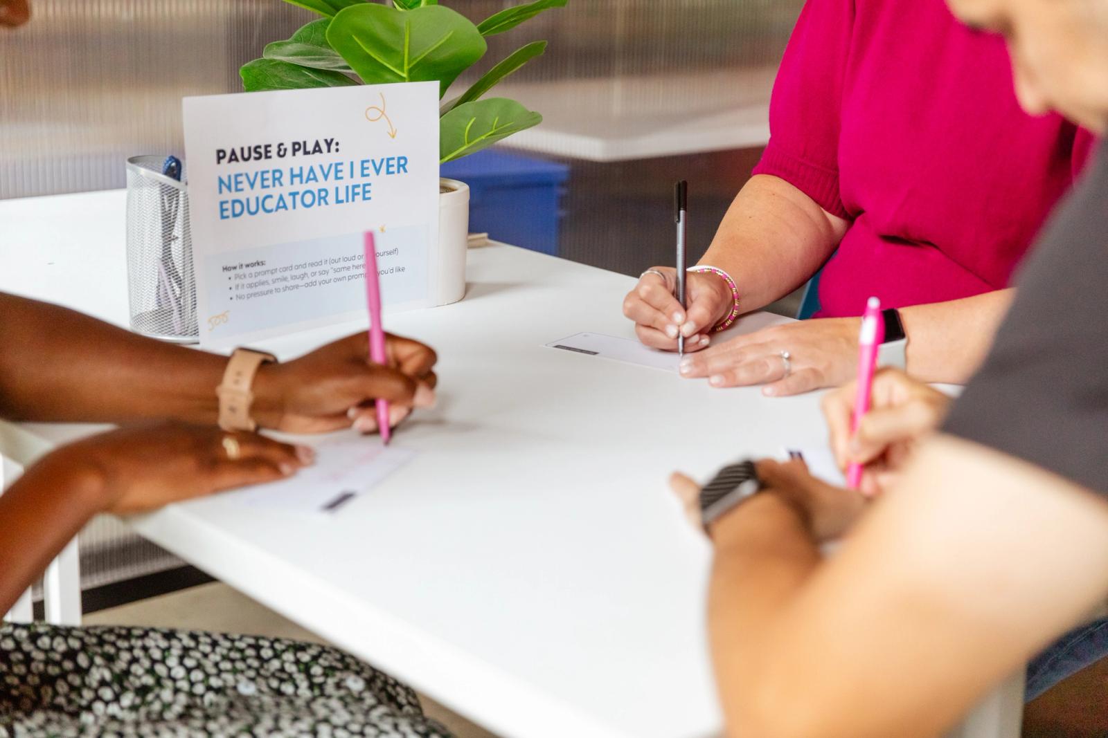Three people sit at a table writing. A piece of paper says "pause & play: never have I ever educator life."
