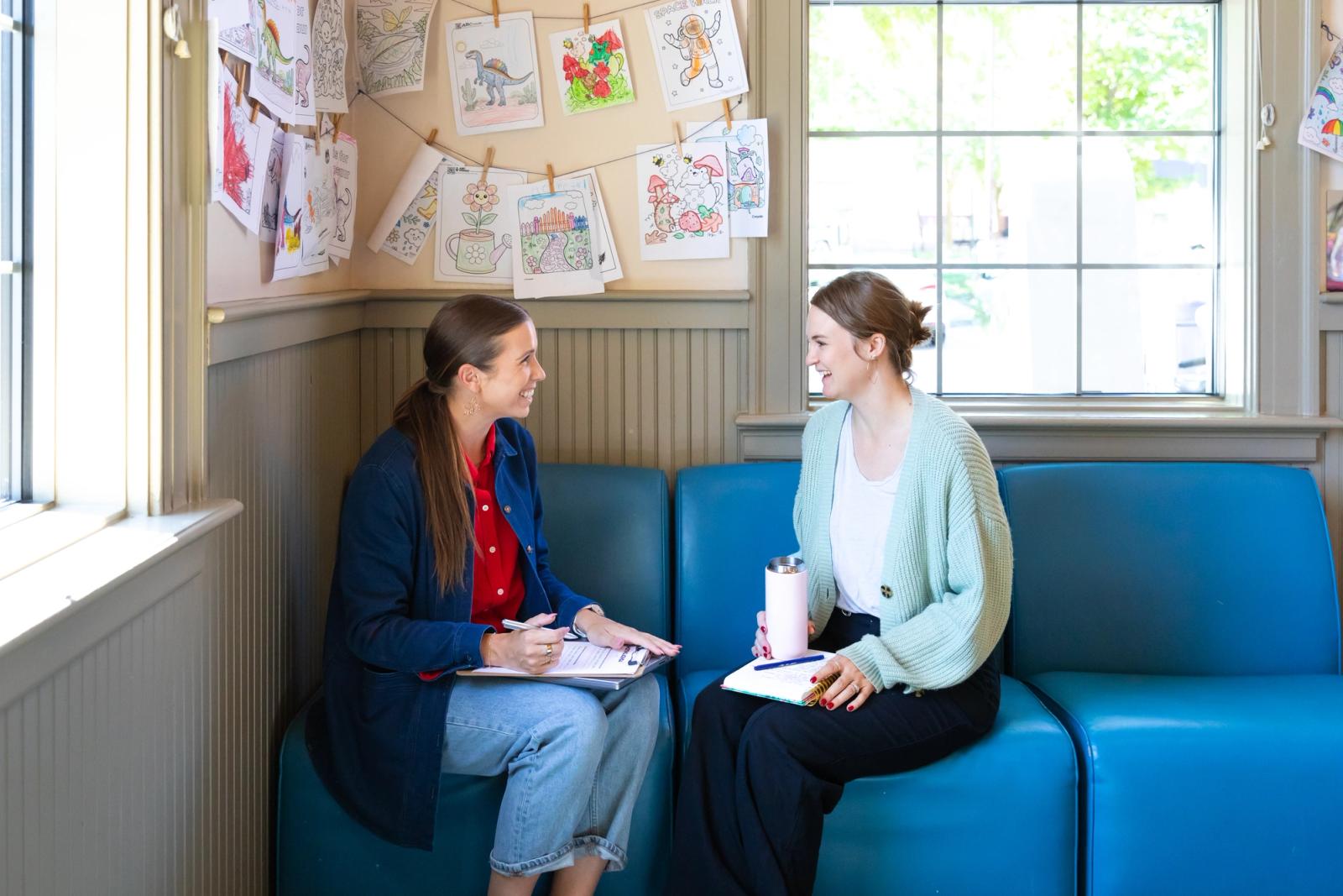 Two women sit on a bench in front of a window and wall with children's drawings on it. They are smiling at each other and have notebooks in the laps.