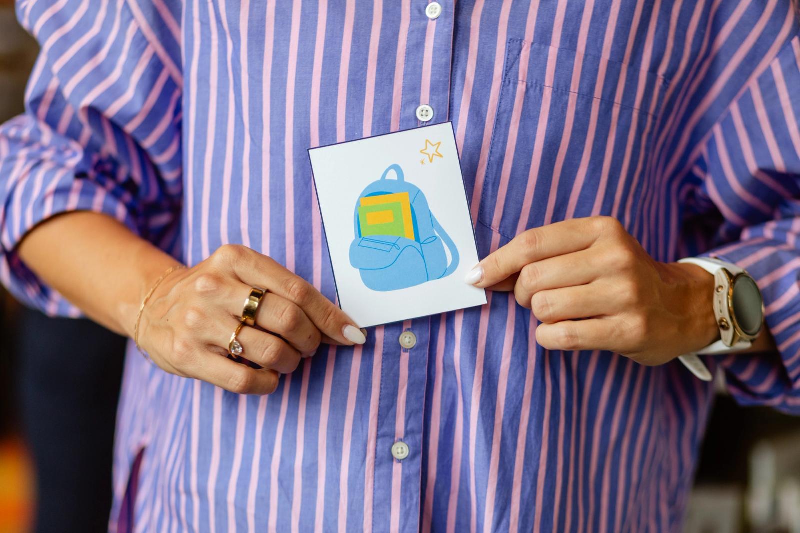 Someone in a blue and pink striped shirt holds a piece of paper with an image of a backpack on it in front of them.