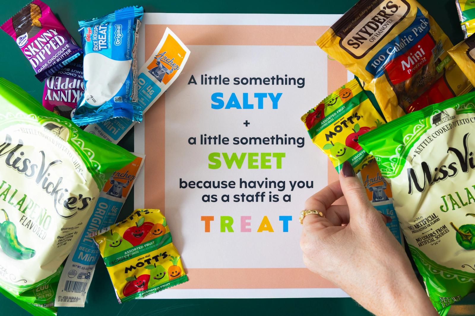 A colorful poster lays on a table. Scattered around it are small snacks, and a hand reaches in to grab a snack.