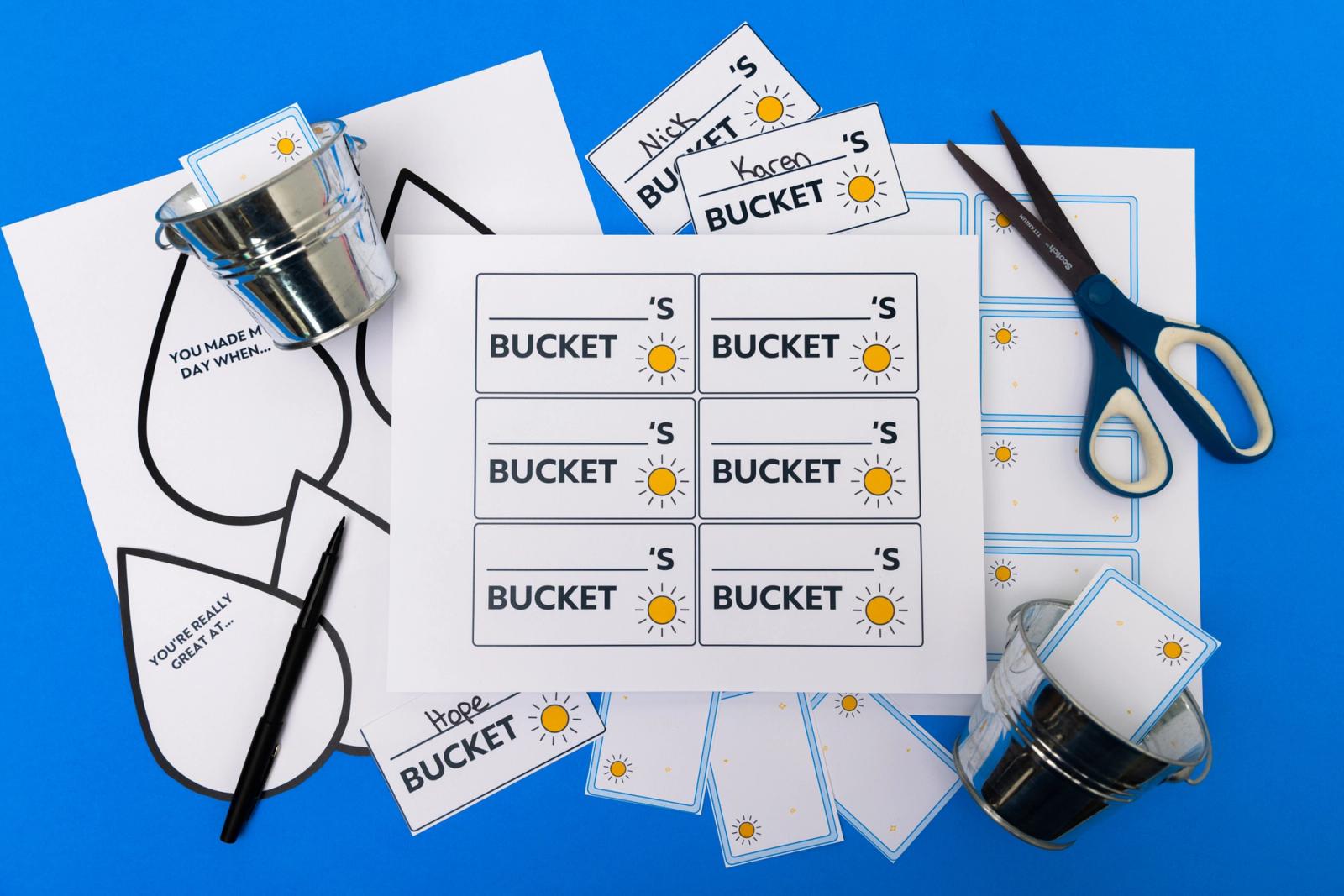 Several sheets of paper, scissors, marker, and small metal pails lay on a blue background.