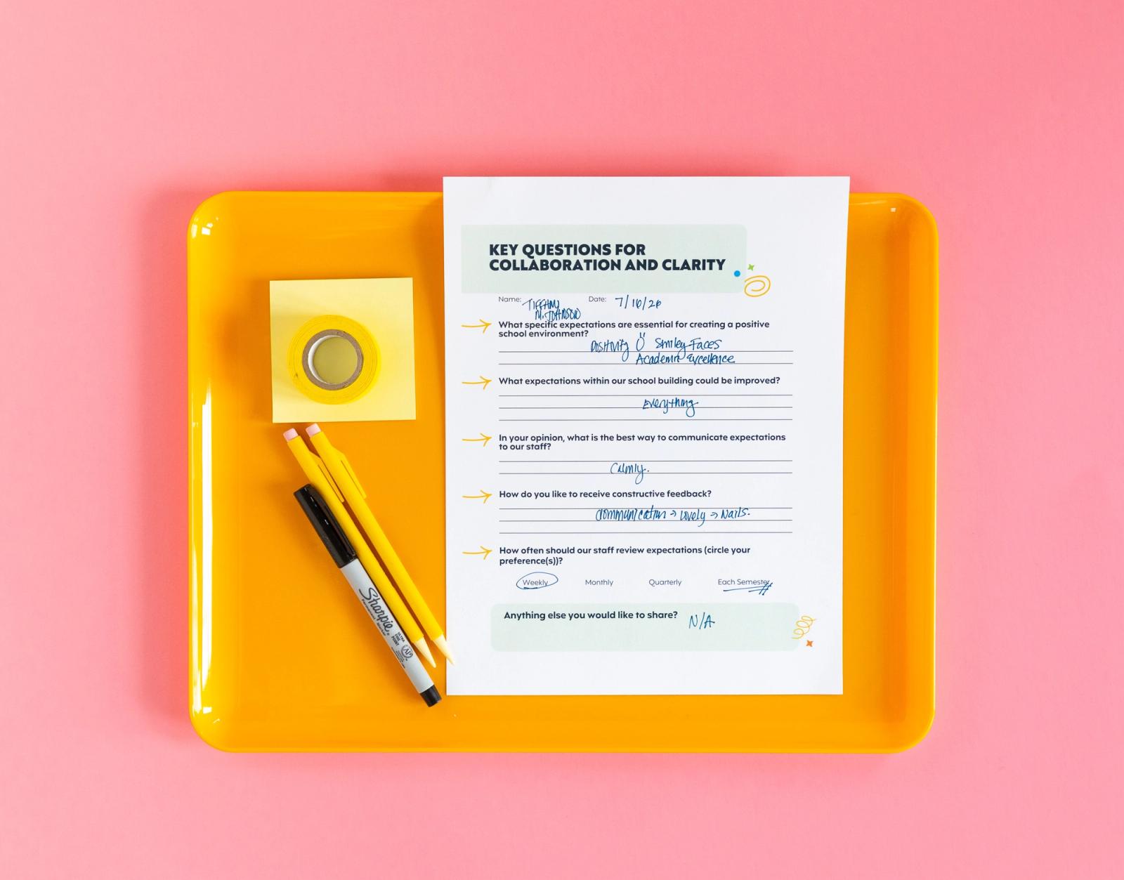 An orange tray with a piece of paper, pencils, marker and sticky notes sits on a pink background.