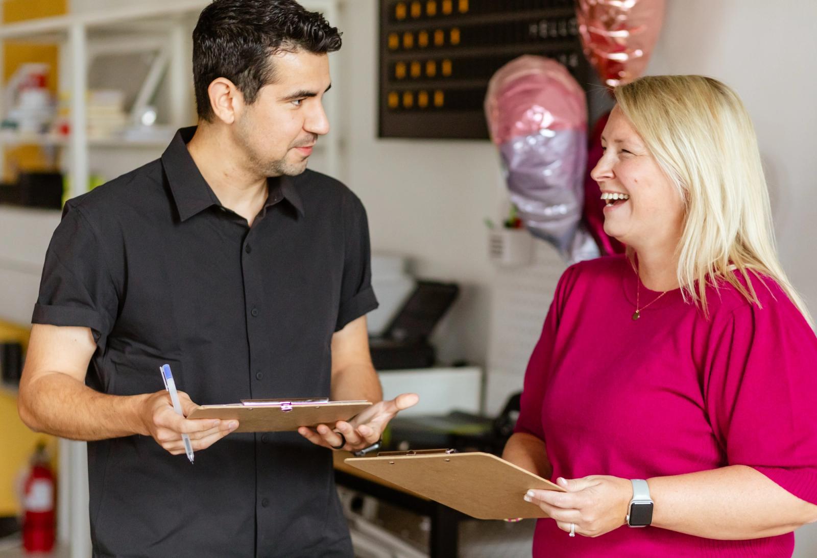 A man and woman stand facing each other. Each holds a clipboard. They are talking and laughing.