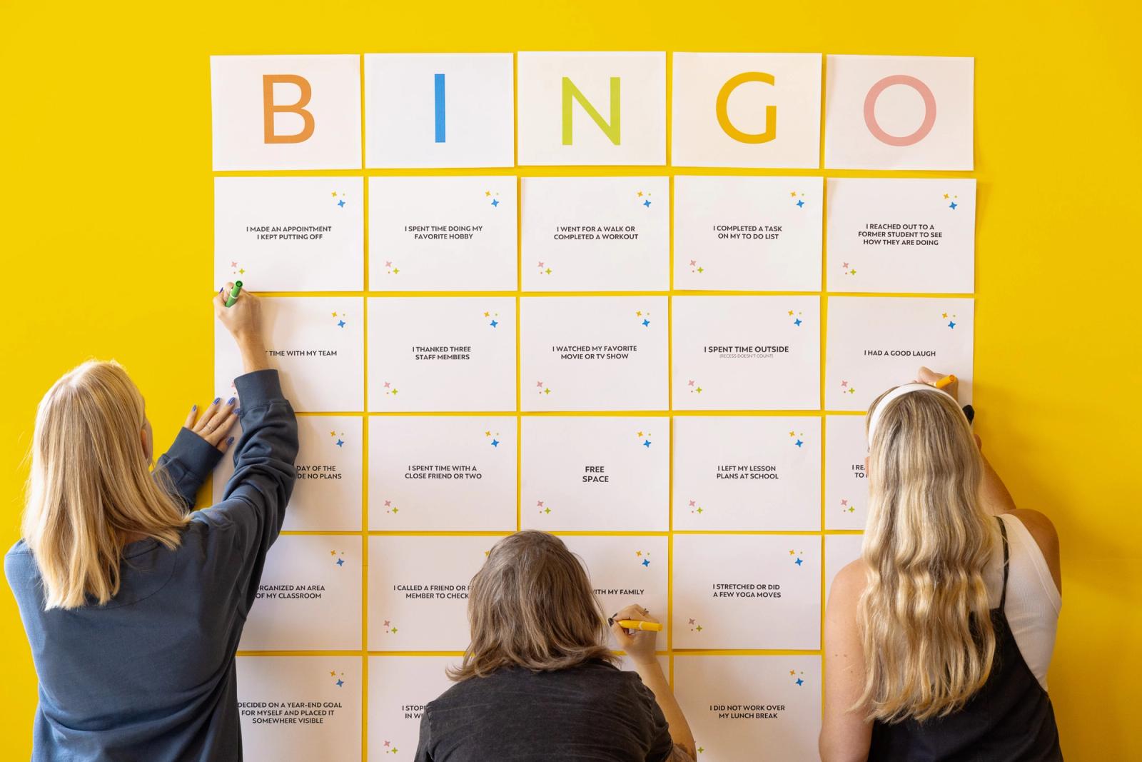 Three women stand in front of a yellow wall with bingo squares on it. They write on the squares.