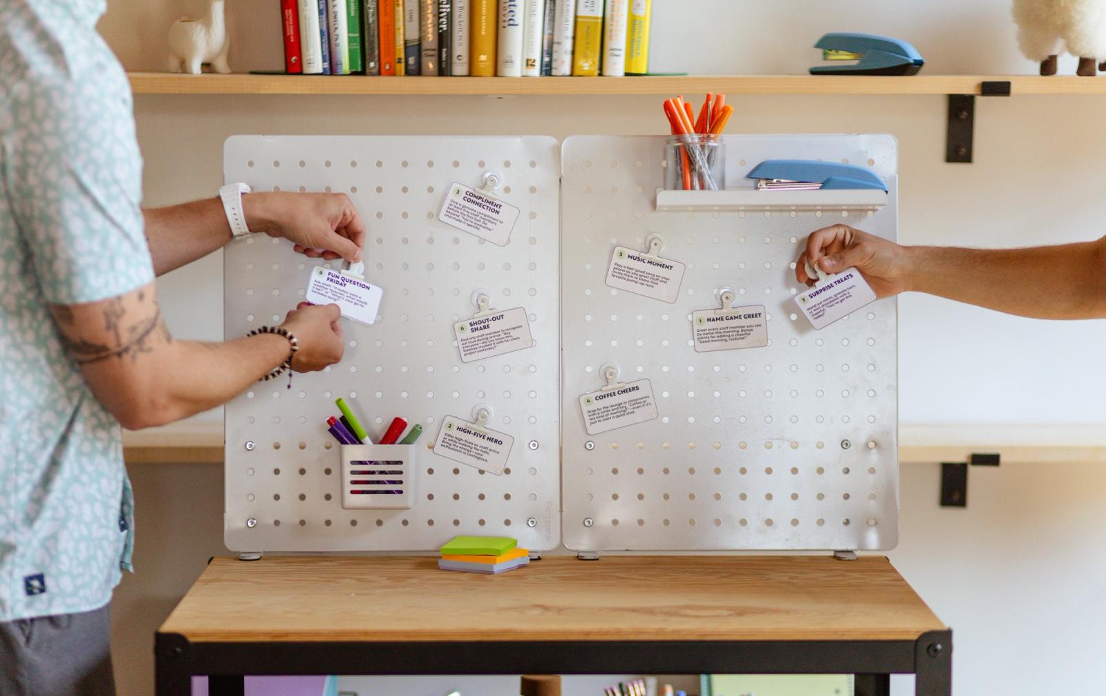 A white peg board sits in front of a white wall with shelves on it. Small notes are attached to the peg board. Two men reach in from either side to place more notes.