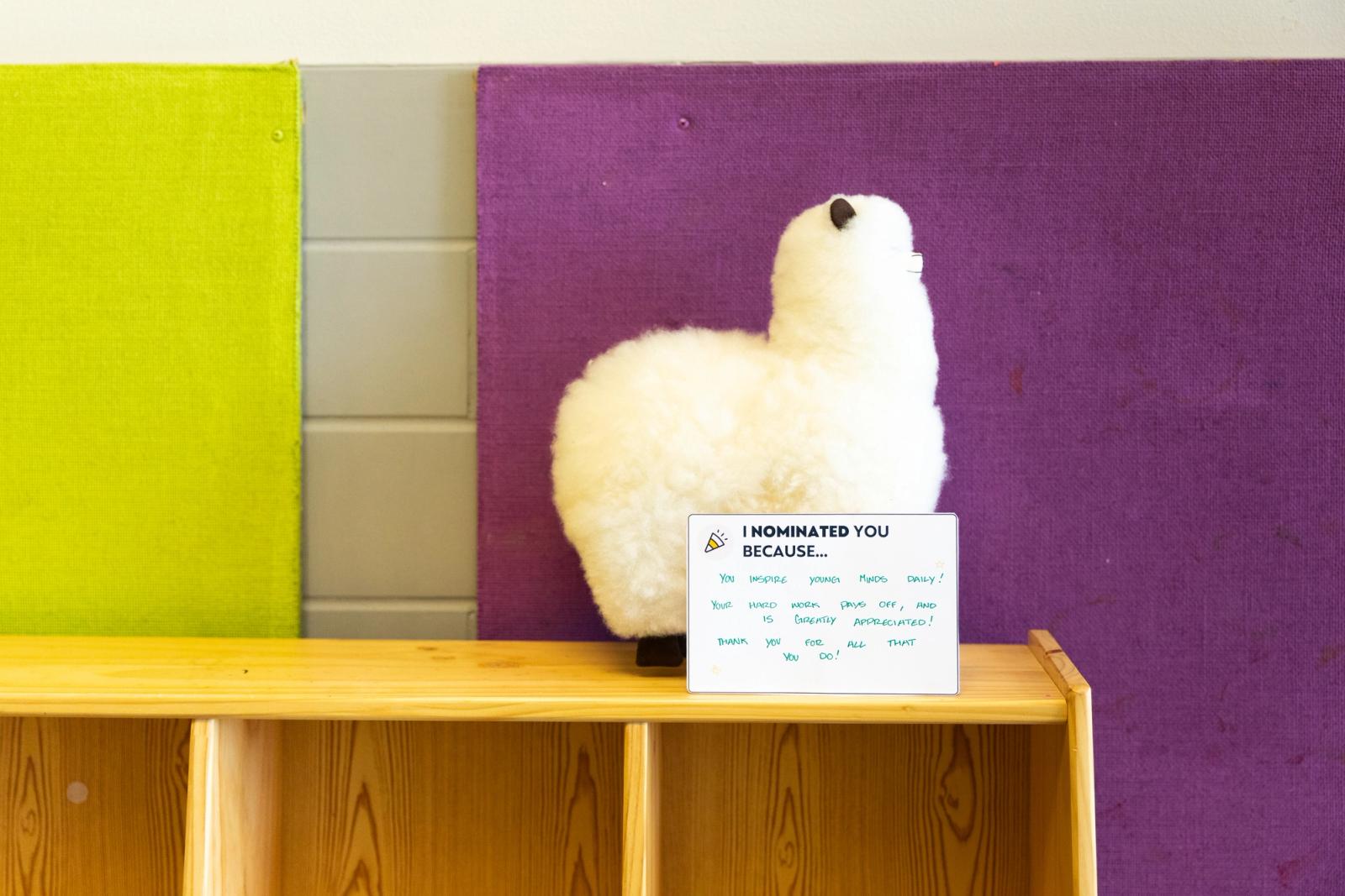 A white, stuffed alpaca sits on a shelf in front of green and purple bulletin boards. A handwritten note is propped up in front of the alapca.