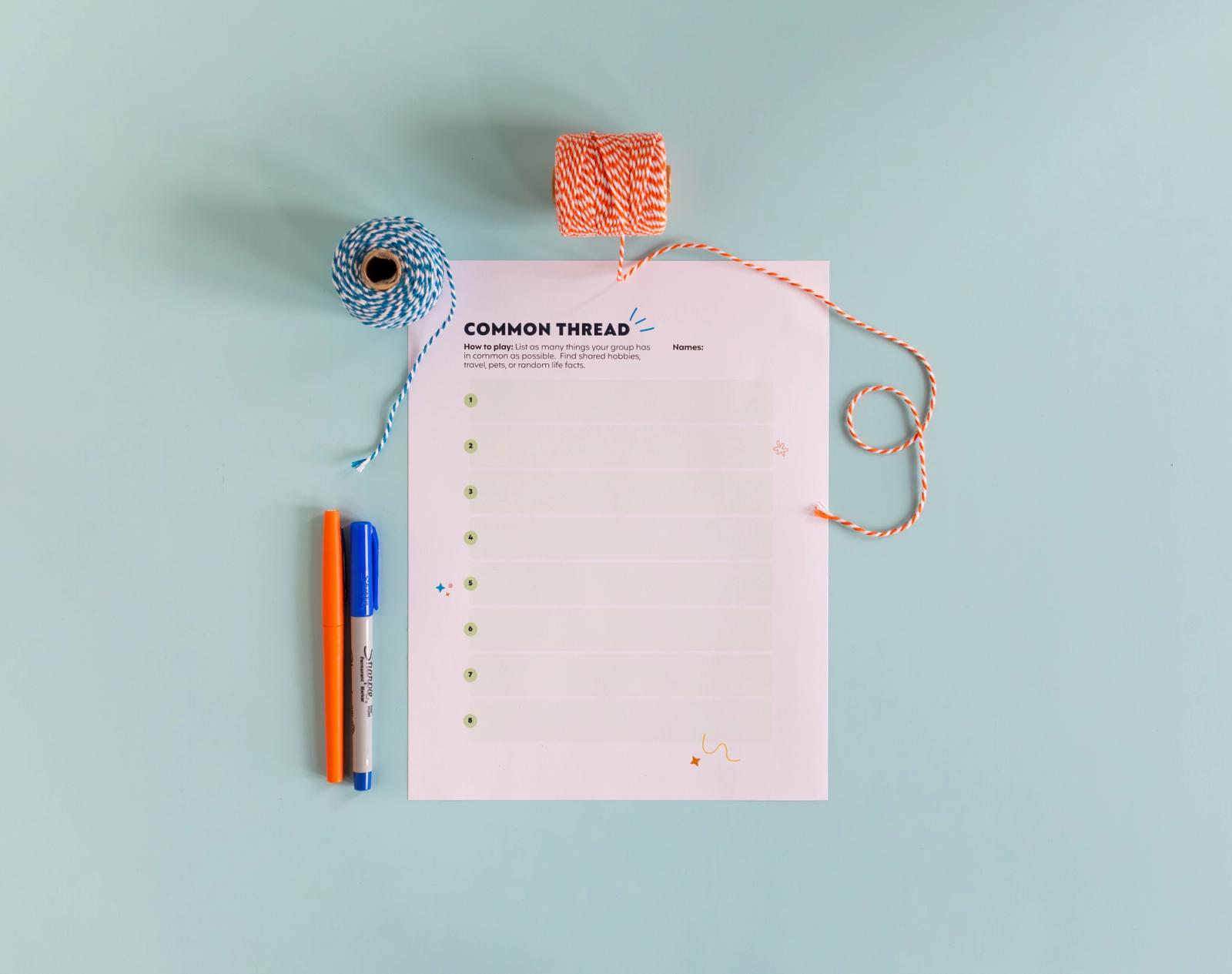 A piece of paper lays on a light blue background. Spools with blue and orange, and orange and blue pens are around the paper.