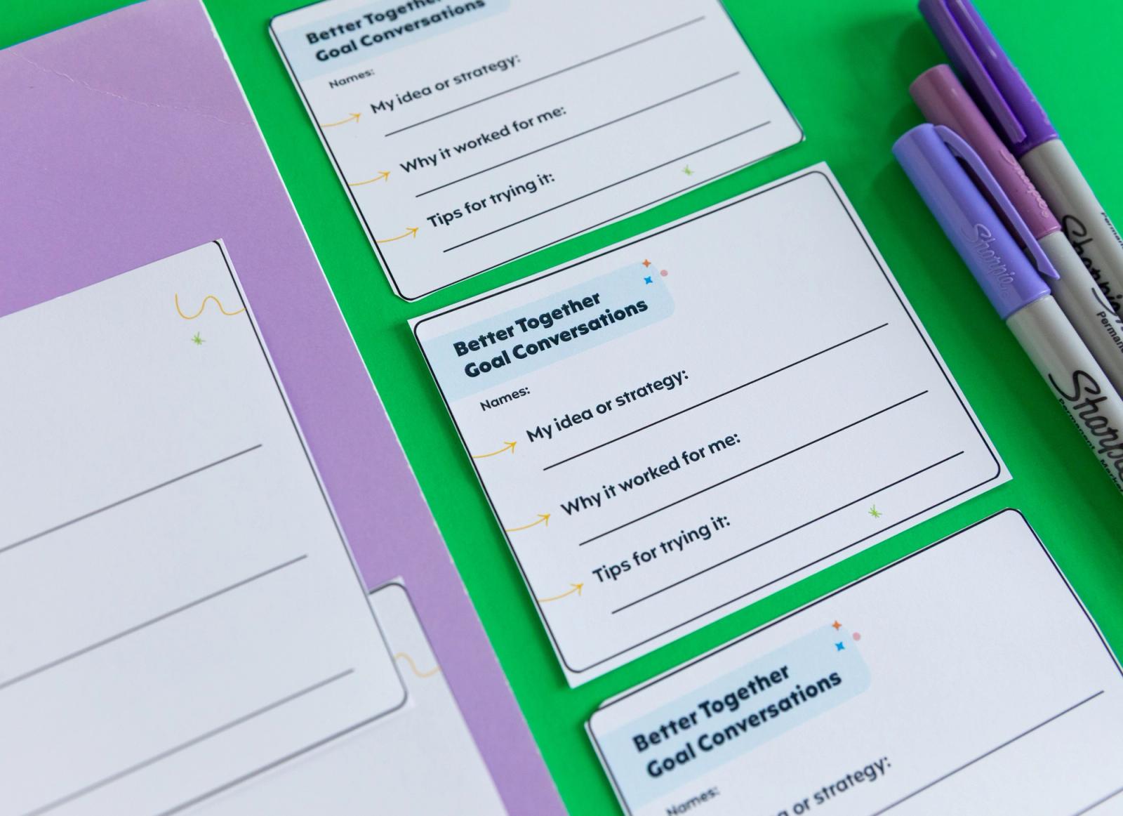 Closeup of three notecards on a green background. Next to it are purple pens and a purple notebook.