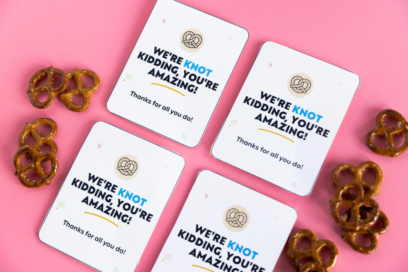 Four small pieces of paper lay diagonally on a pink background. Pretzels lay around them.