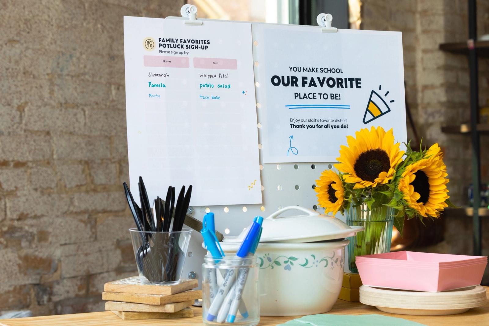 A casserole dish, utensils, plates, pens and sunflowers sit on a table in front of a brick wall. Two posters are handing on the wall.