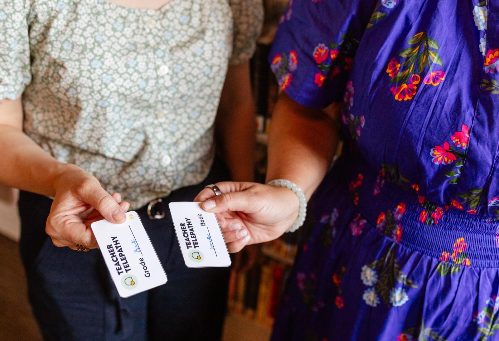 Two women stand and each hold a small piece of paper toward one another.