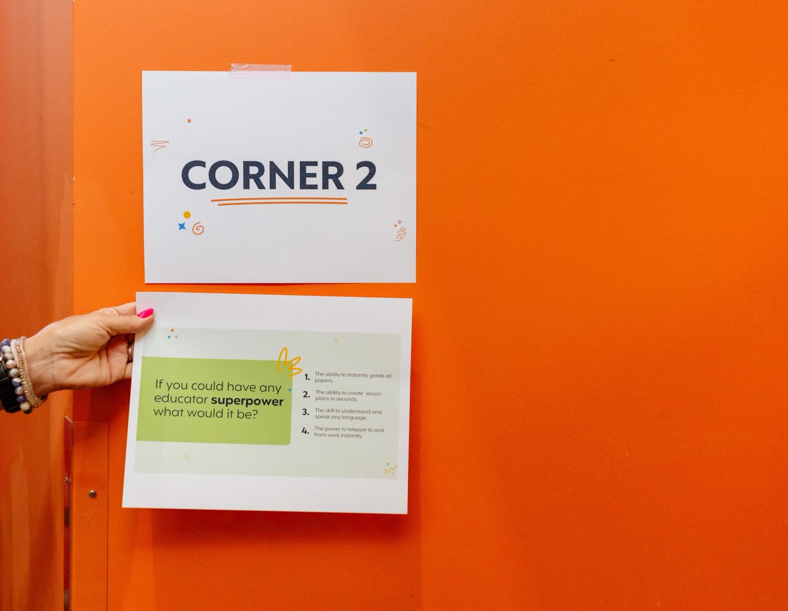 A poster that says "corner 2" is taped to an orange wall. A hand reaches in to place a list of questions below it.