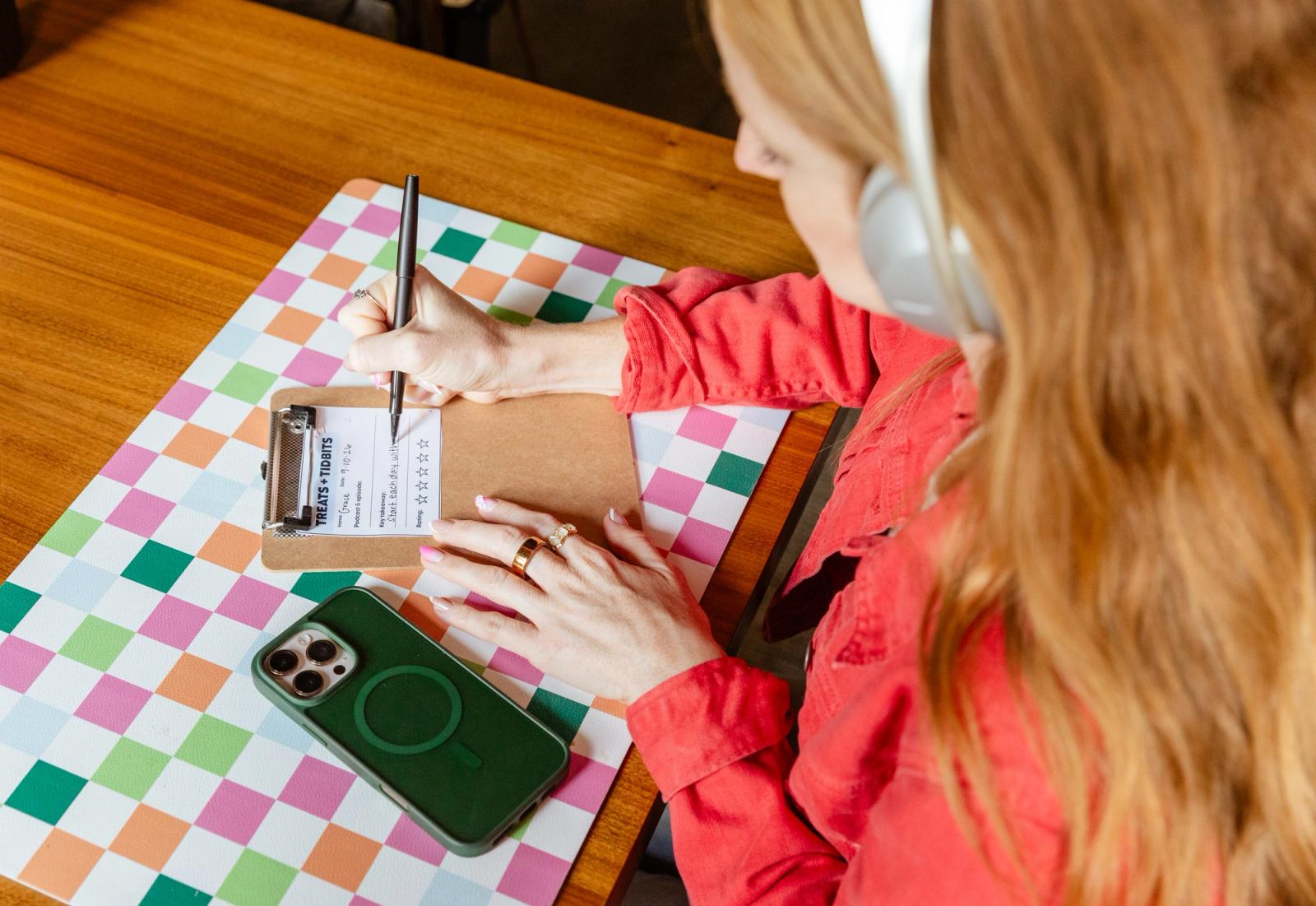 A woman sits at a desk with headphones on. She writes on a piece of paper on a clipboard.