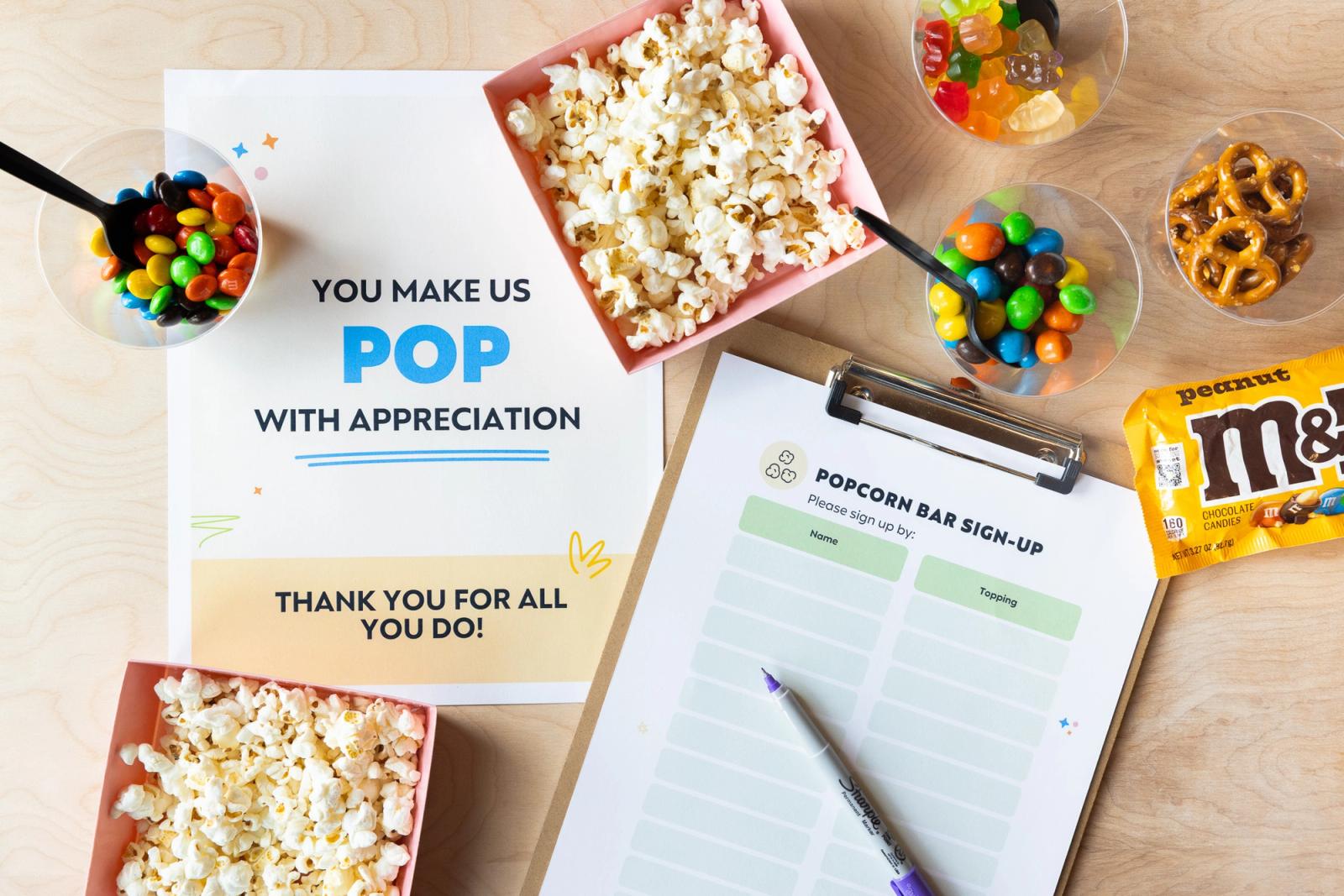 A poster, clipboard with a sheet of paper, and containers of popcorn lay on a table. Scattered around are candies and snacks. 