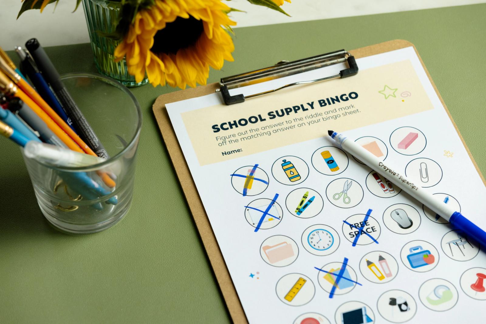 A clipboard with a school supply bingo sheet on it, and a jar of pencils lay on a desk with flowers.