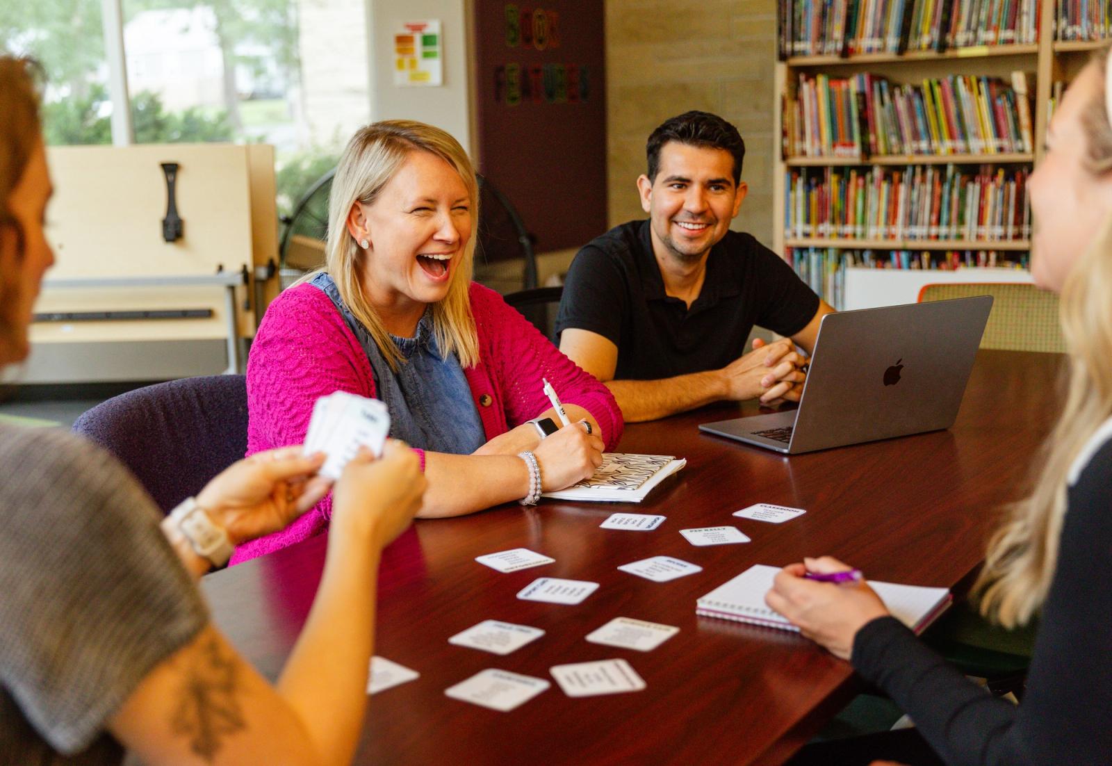 A group of three women and one man sit at a table in a library playing a card game.