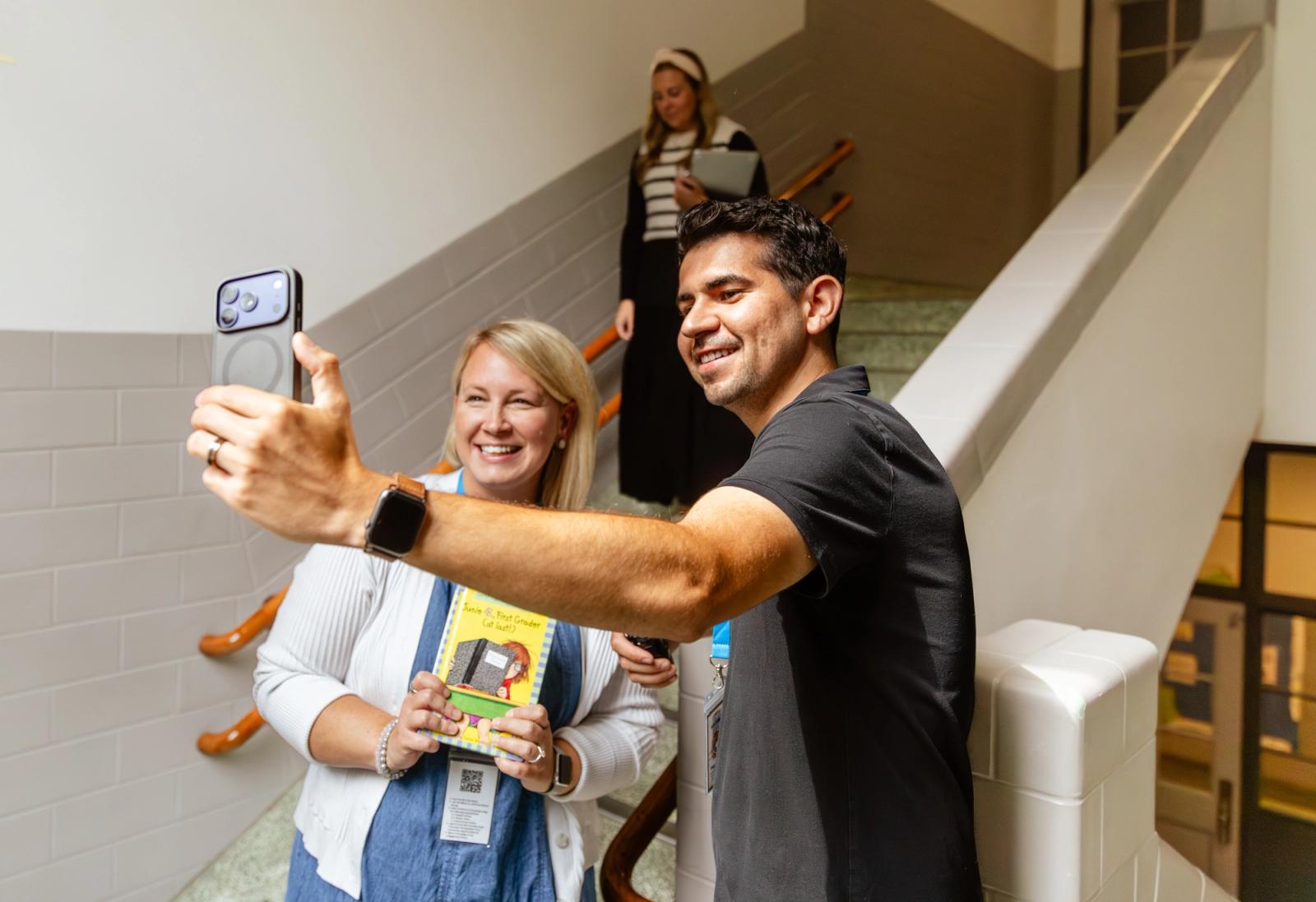 A man and a woman take a selfie in a stairwell. A woman goes down the stairs behind them. 